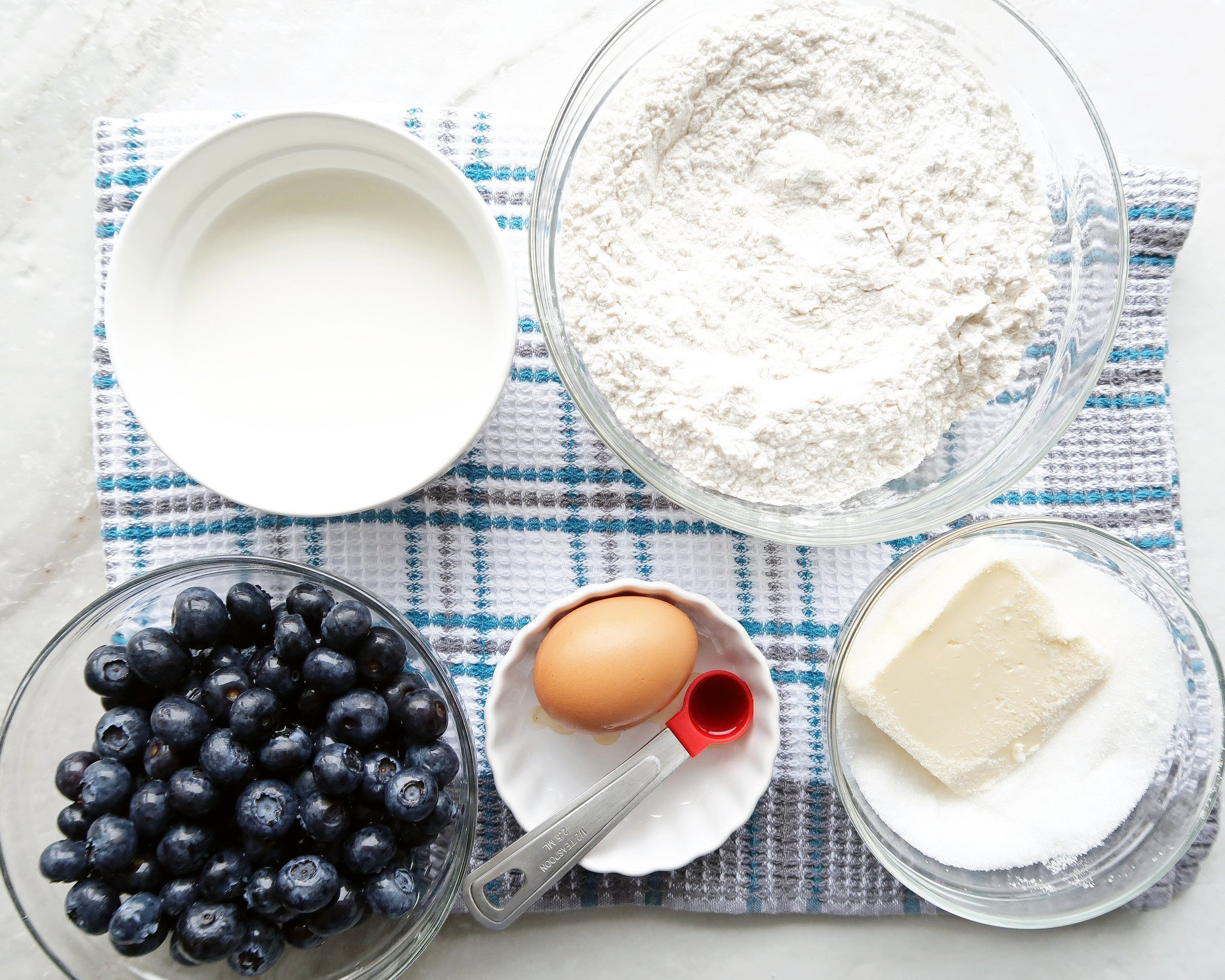 Many bowls containing blueberries, an egg, butter, flour, baking powder, sugar, vanilla,and milk.
