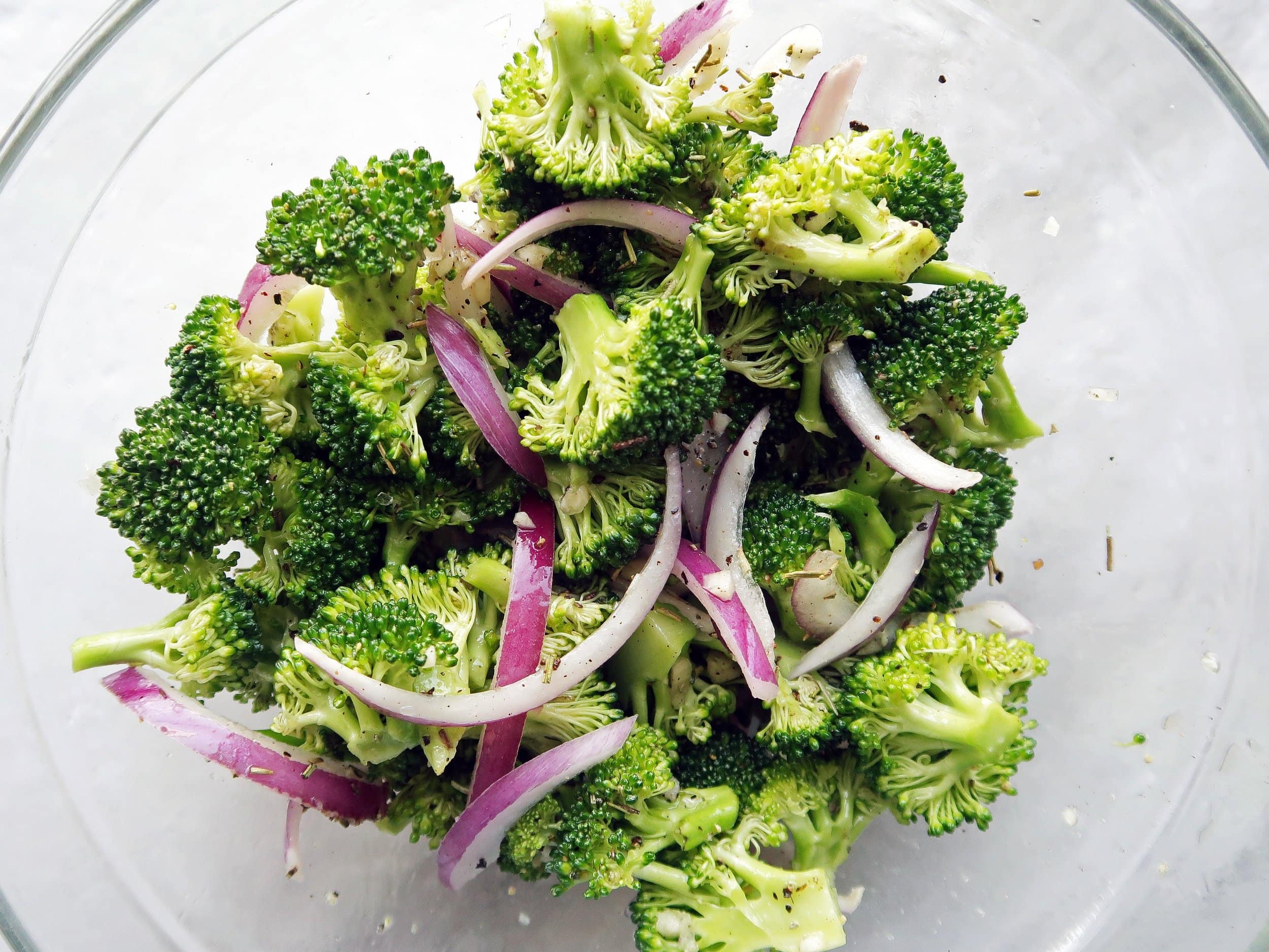 Broccoli, red onion slices, garlic, and herbs in a glass bowl.
