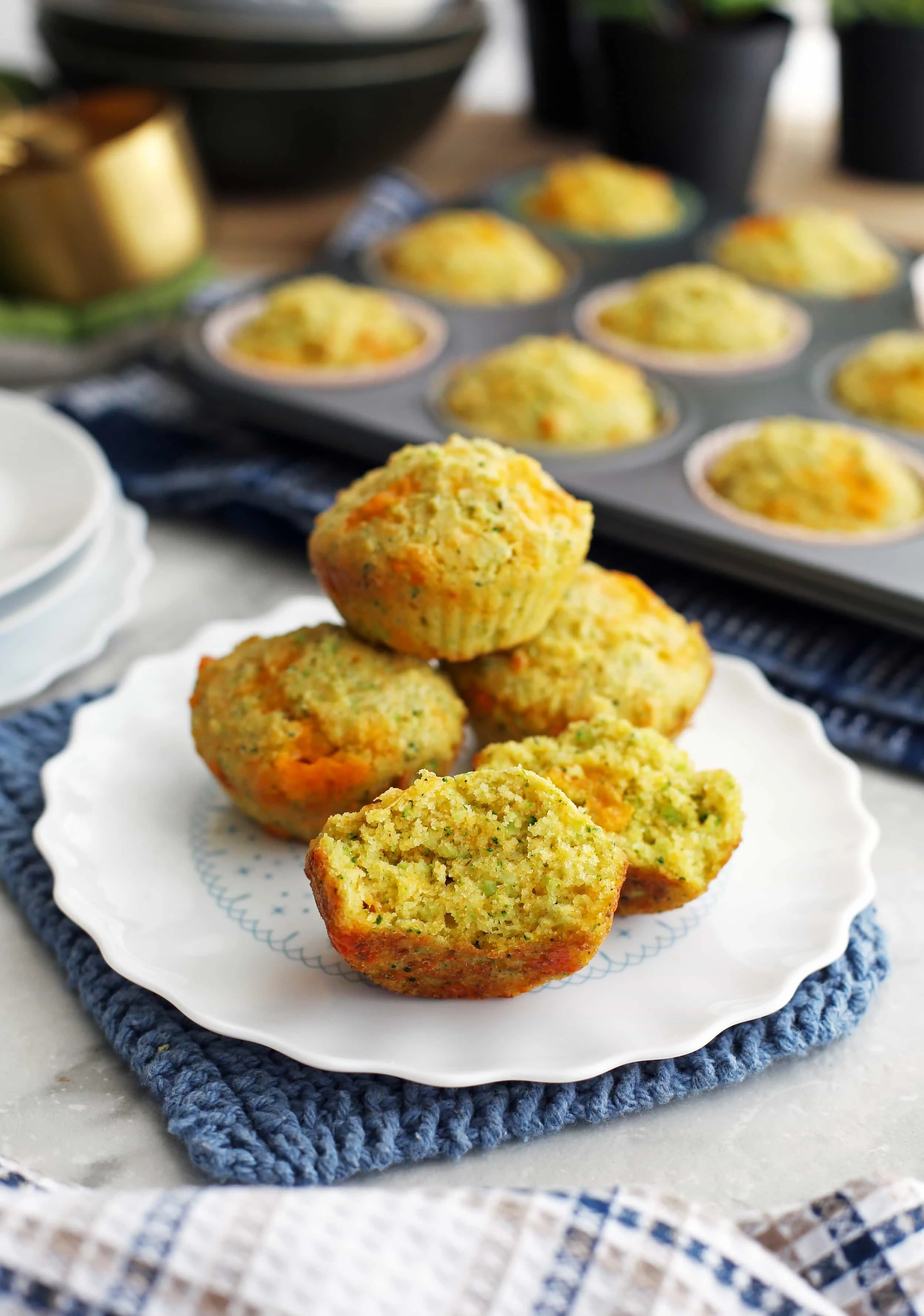 Four Broccoli Cheddar Cornbread Muffins with one cut in half on a white plate with more in a muffin pan behind them.