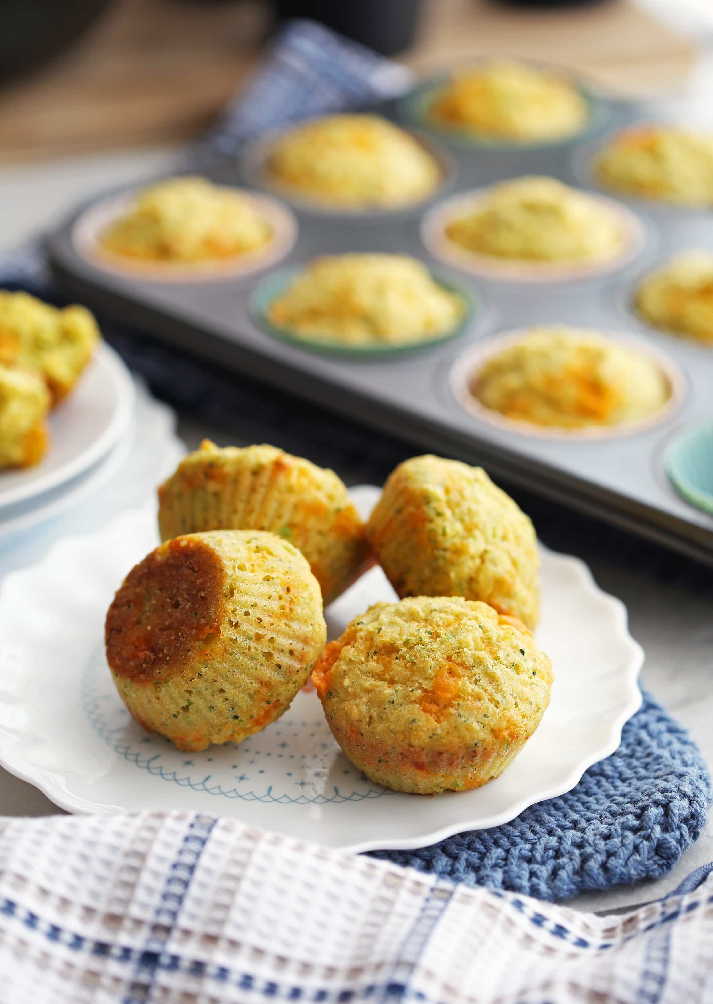 Four Broccoli Cheddar Cornbread Muffins on a white plate with more muffins in a muffin pan behind them.