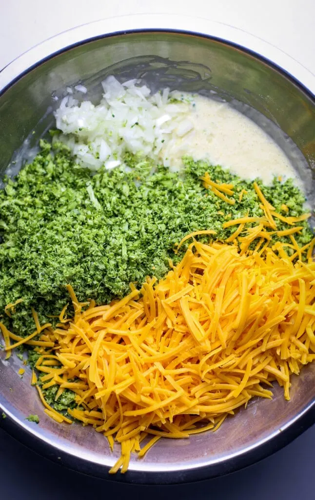 Grated cheddar cheese, broccoli rice, diced onion, and flour mixture in stainless steel bowl.