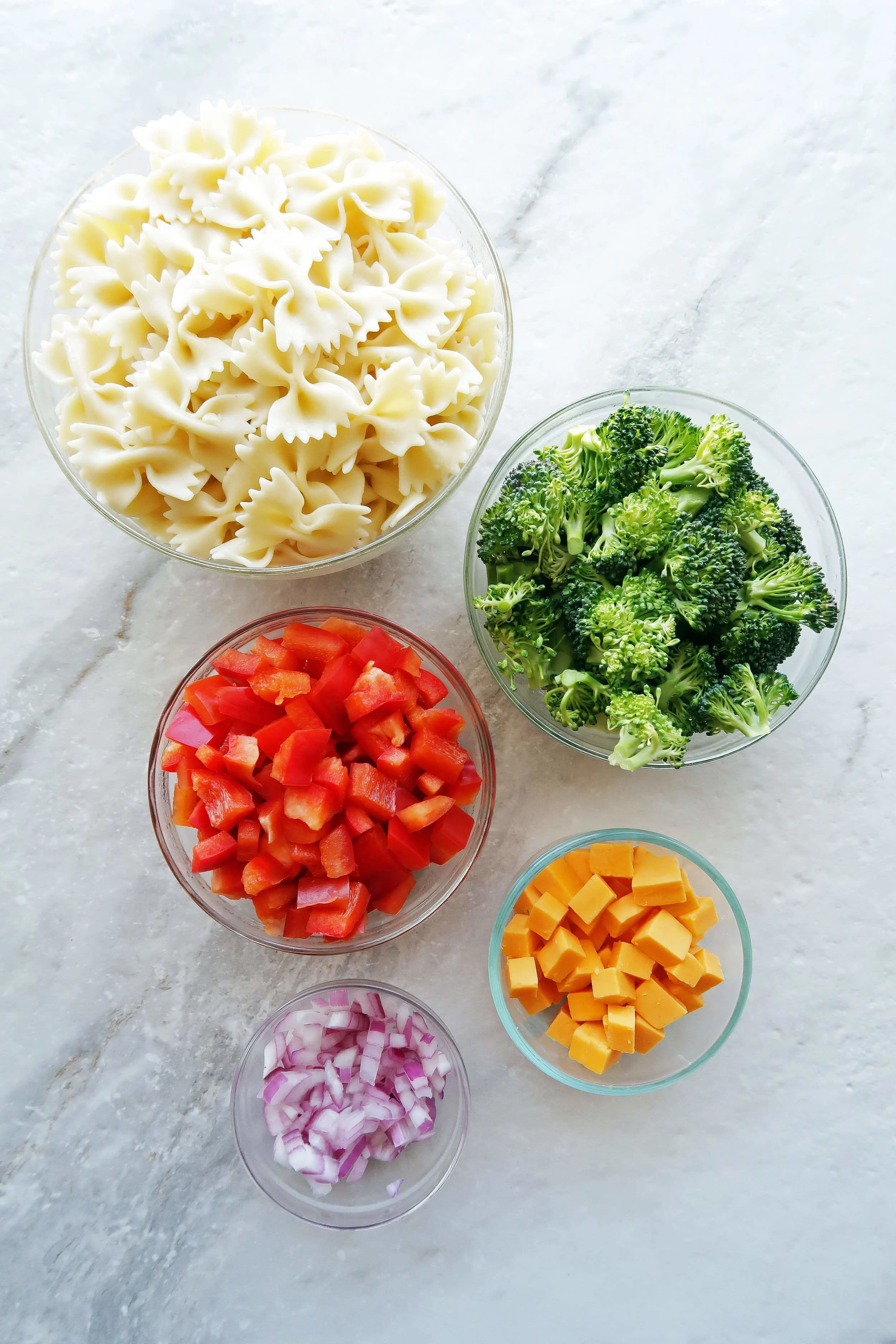 Bowls of bowtie pasta, broccoli, red bell peppers, cheddar cheese, and red onions.