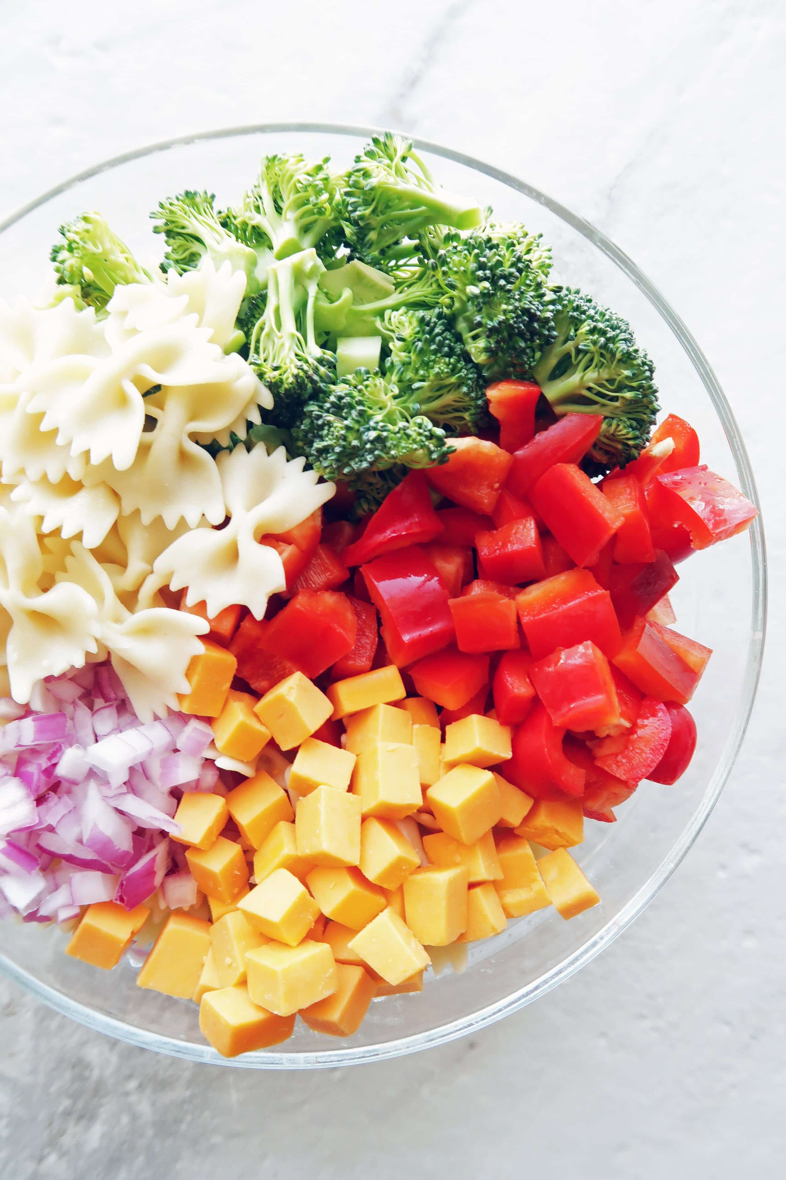 A big bowl of bowtie pasta, broccoli, red bell peppers, cheddar cheese, and red onions.