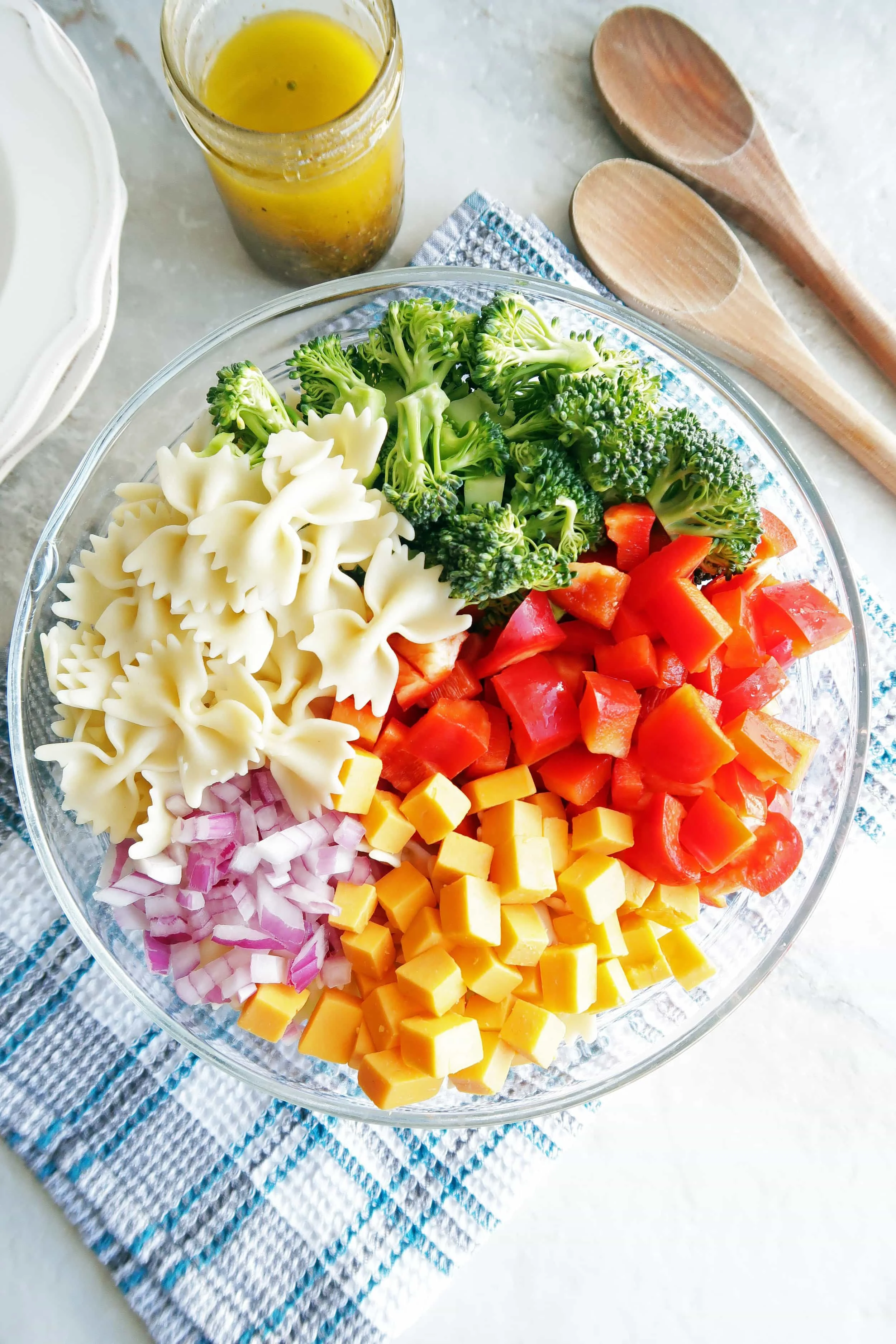  Unmixed Broccoli Cheddar Pasta Salad in a large glass bowl with Italian Vinaigrette in a jar and two wooden spoons in the background.