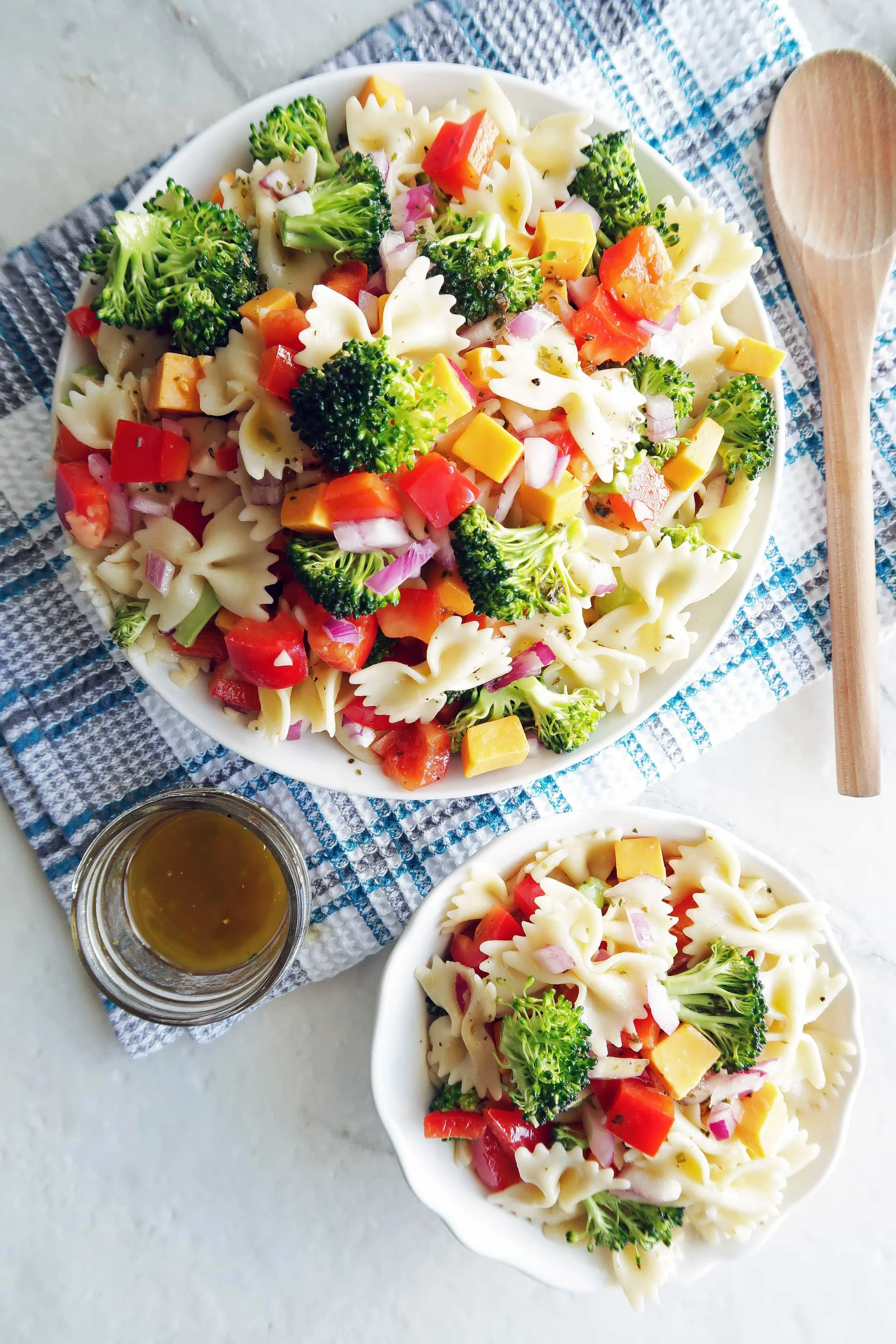 Broccoli Cheddar Pasta Salad with Tangy Italian Vinaigrette in a large pasta bowl, a small bowl of pasta salad, dressing in a small jar, and wooden spoon to the side.