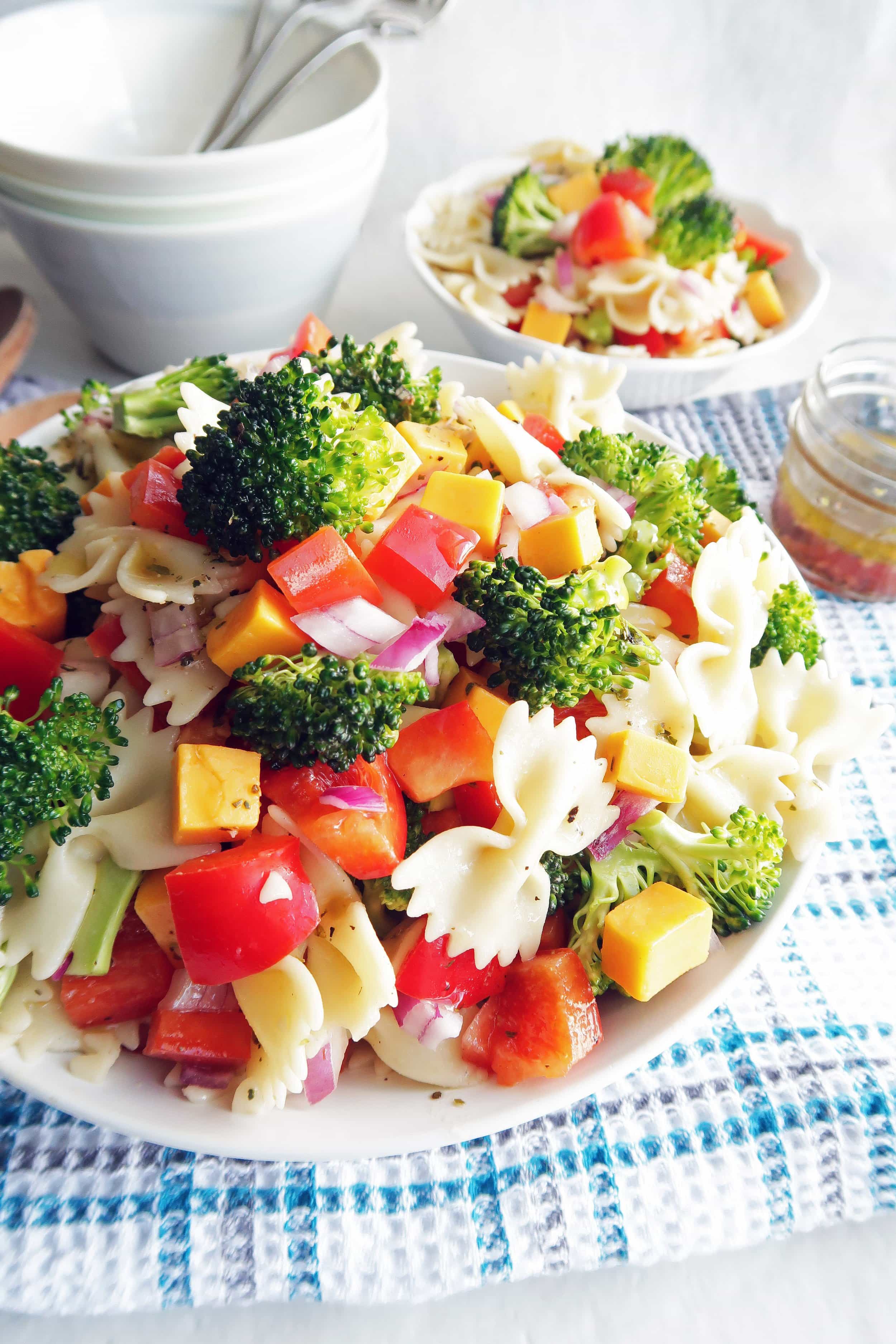 Side view of broccoli Cheddar Pasta Salad with Tangy Italian Vinaigrette in a large pasta bowl, a small bowl of pasta salad, dressing in a small jar, and bowls and forks in the background.