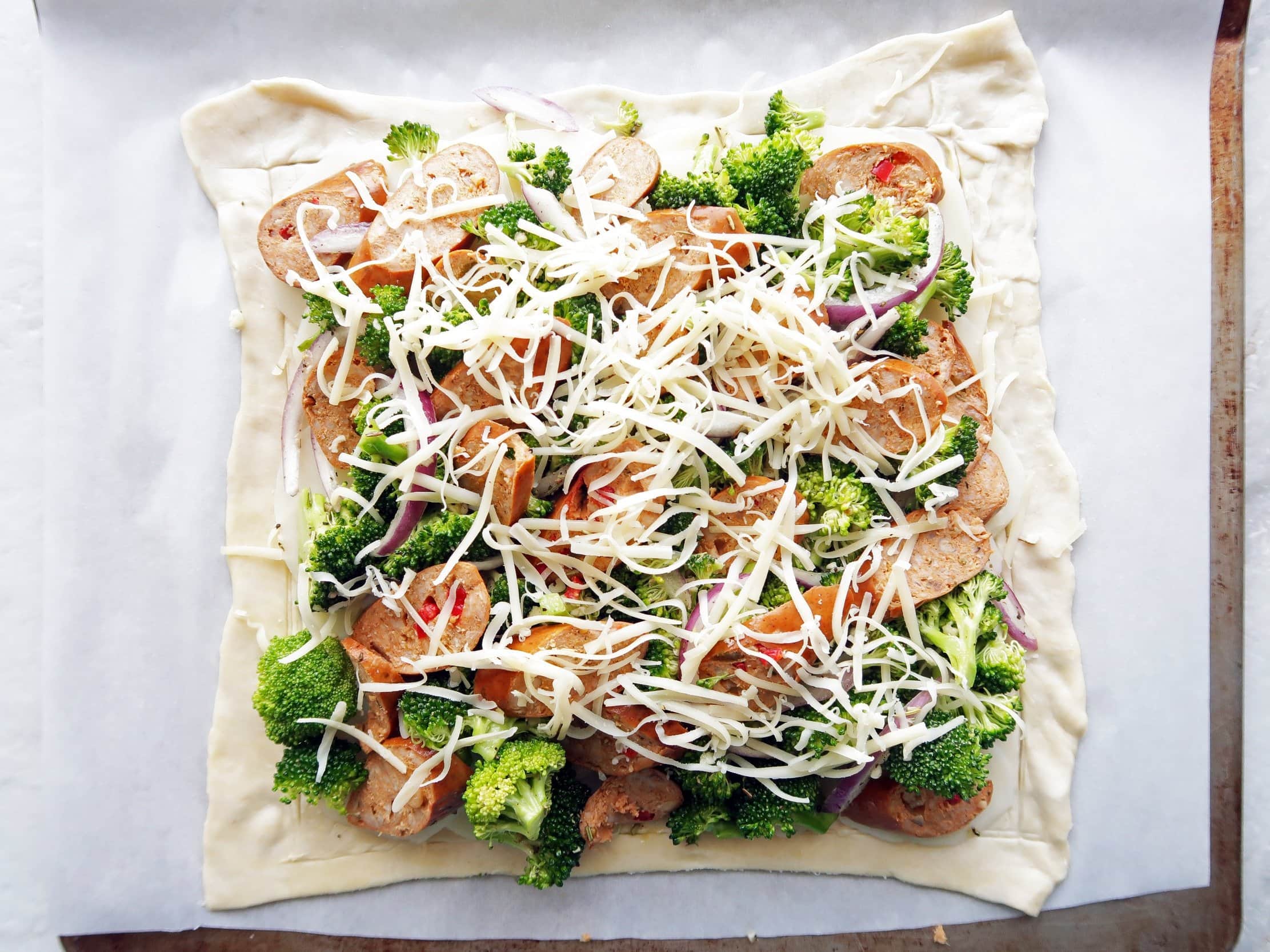 A sheet of puff pastry covered with potatoes, broccoli, sausage, and cheese.