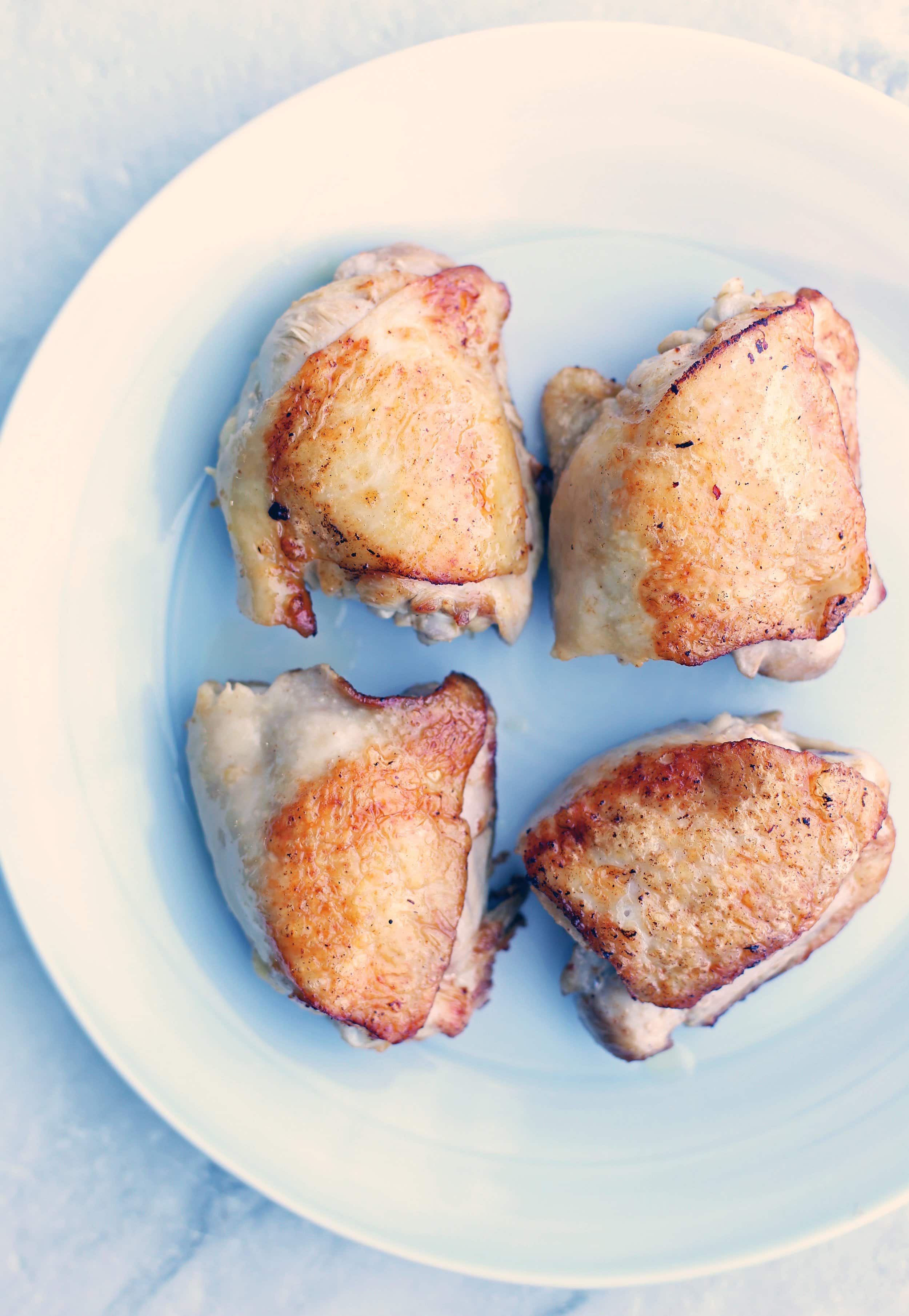 Four bone-in chicken thighs with seared golden-brown skin on a plate.