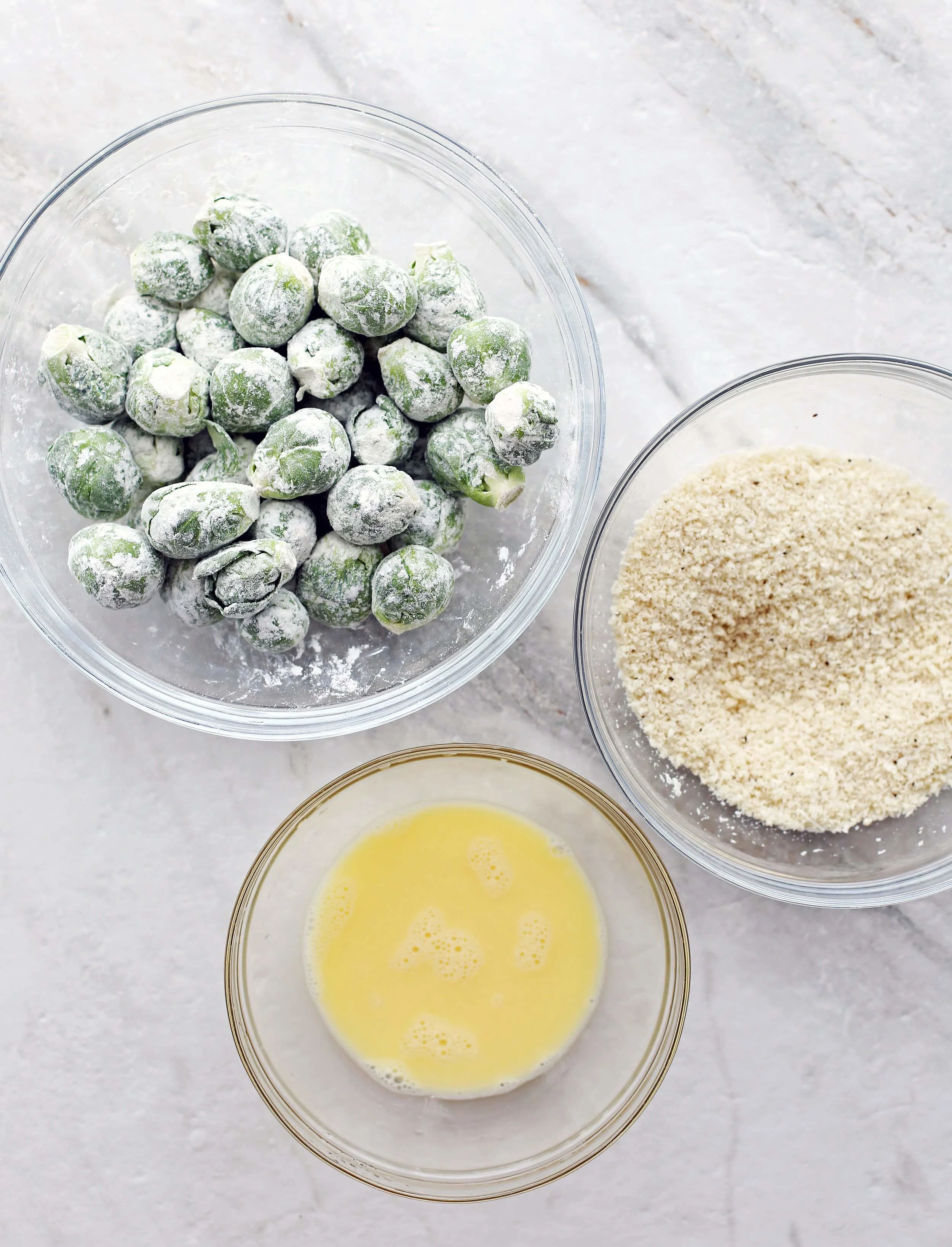Bowls containing beaten eggs, breadcrumbs, and floured Brussels sprouts.