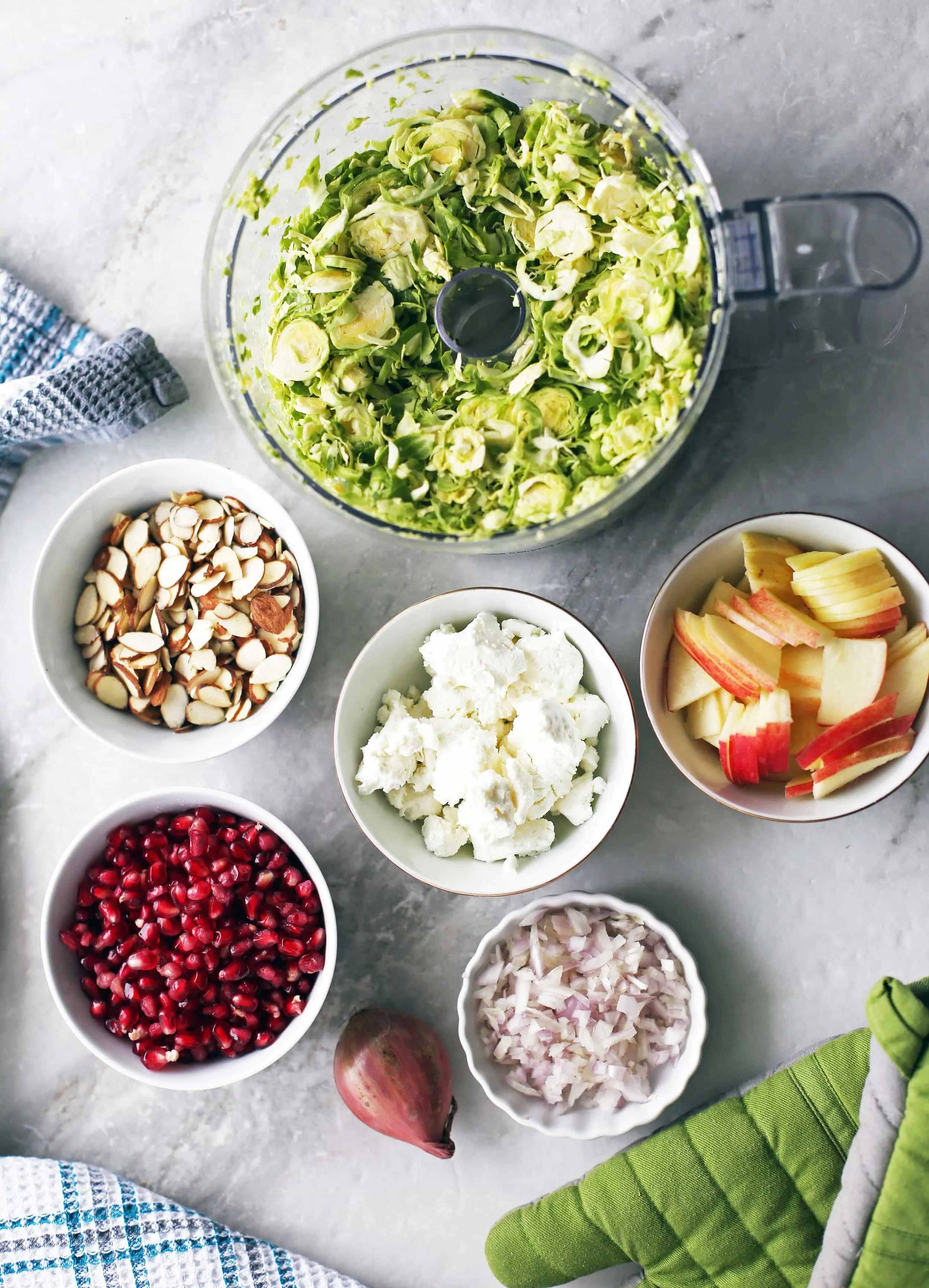 An overhead view of a bowls of shaved Brussels sprouts, almonds, pomegranate arils, shallot, goat cheese, and apple.