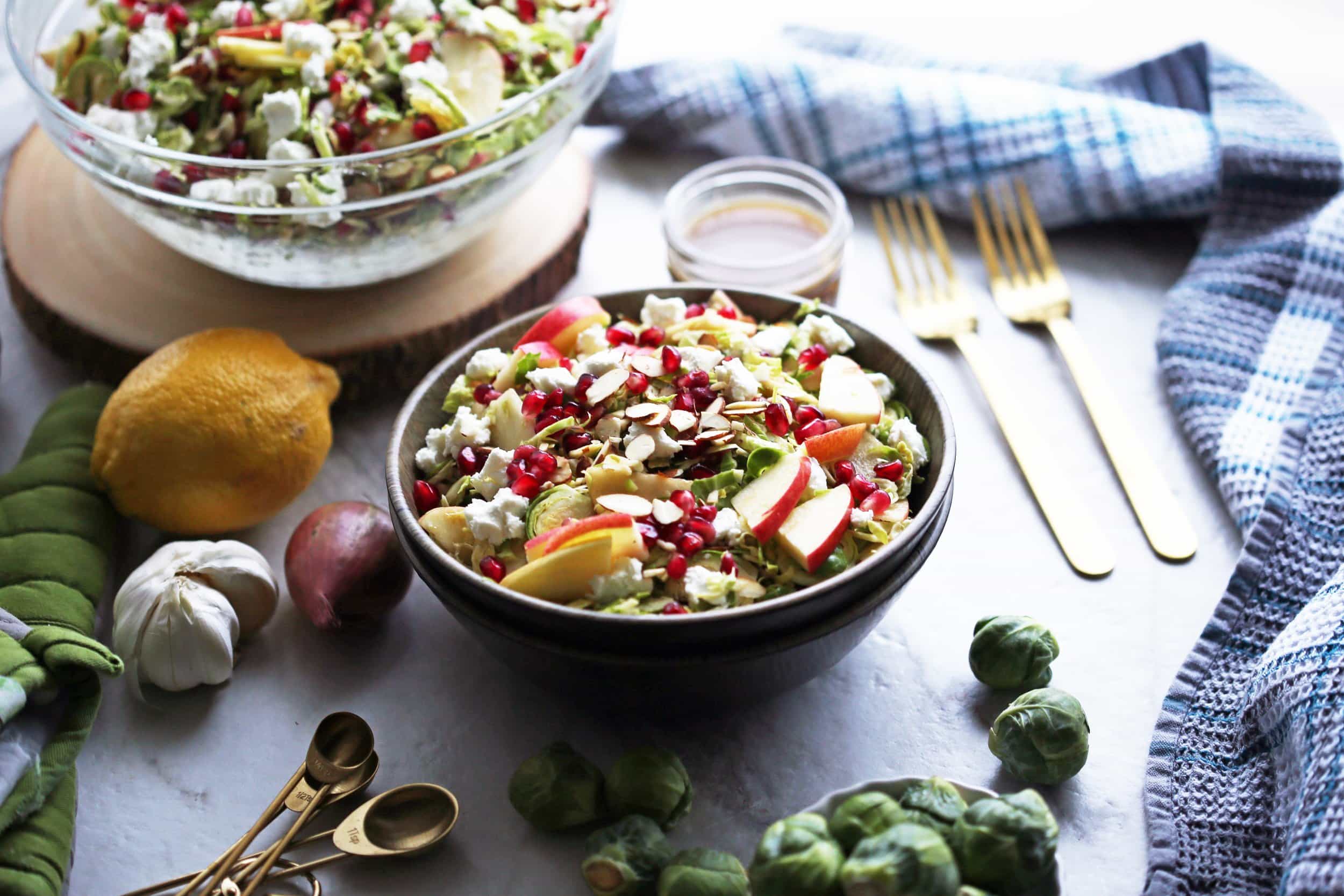 A side angled view of a bowl of shaved Brussels sprouts and pomegranate salad with lemon vinaigrette with more salad in a large glass bowl in the background.