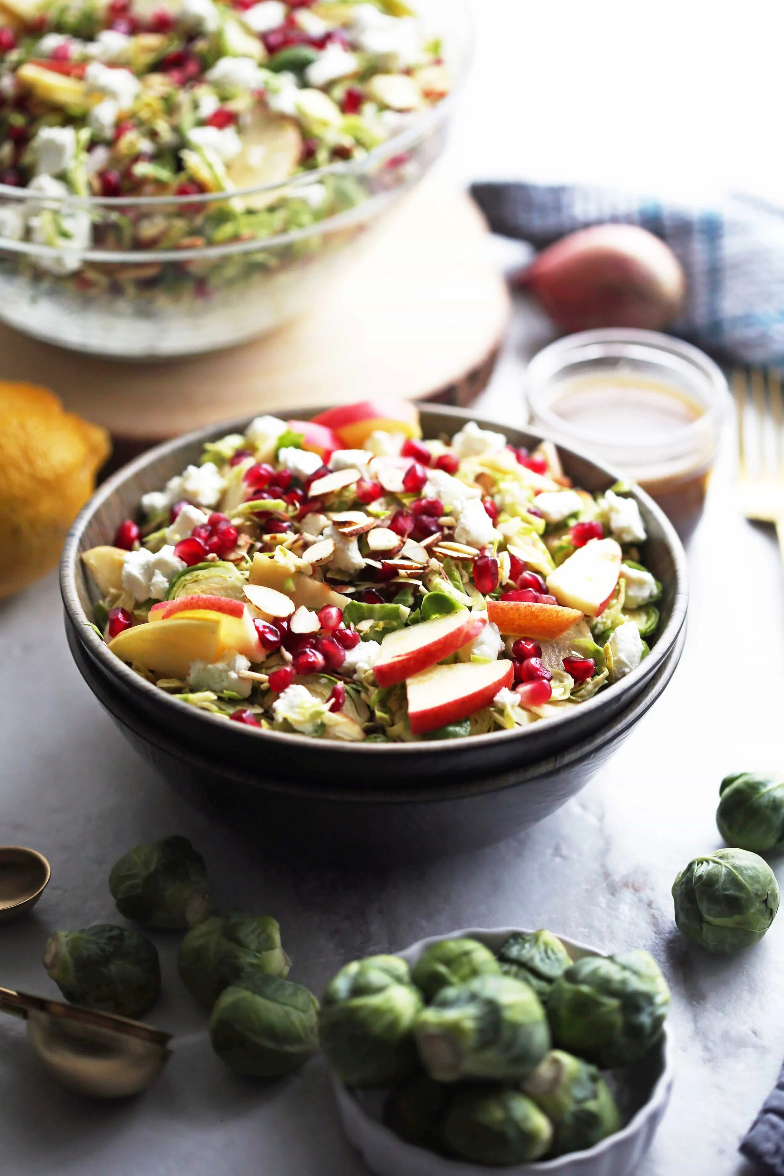 A bowl of shaved Brussels sprouts and pomegranate salad with lemon vinaigrette with more salad in a large glass bowl in the background and raw Brussels sprouts in the foreground.