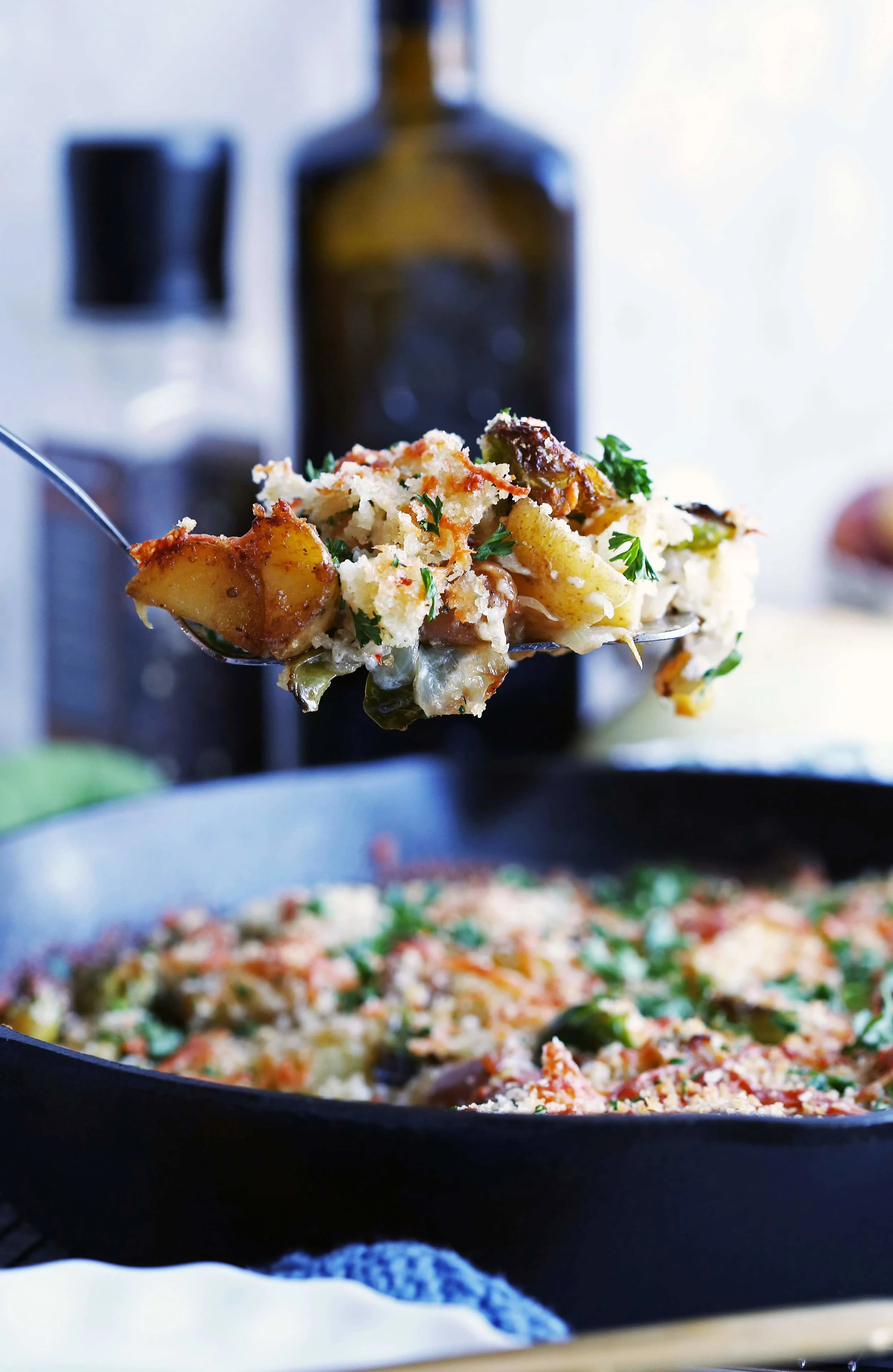 A heaping scoop of Brussels Sprouts and Potato Gratin on a metal spatula held over a large cast iron skillet with more gratin in it.