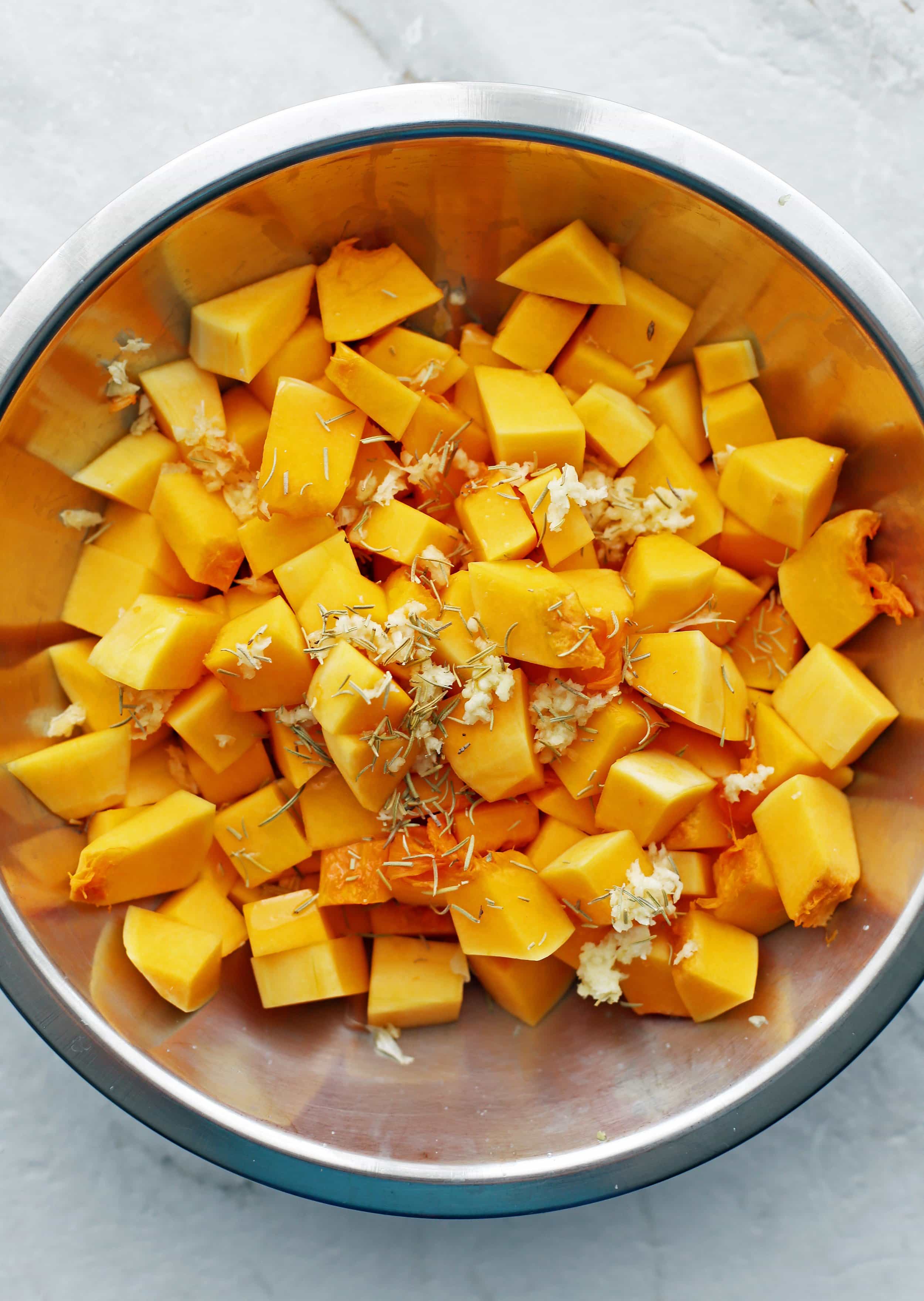 A metal bowl containing chopped butternut squash, garlic, herbs, and olive oil.