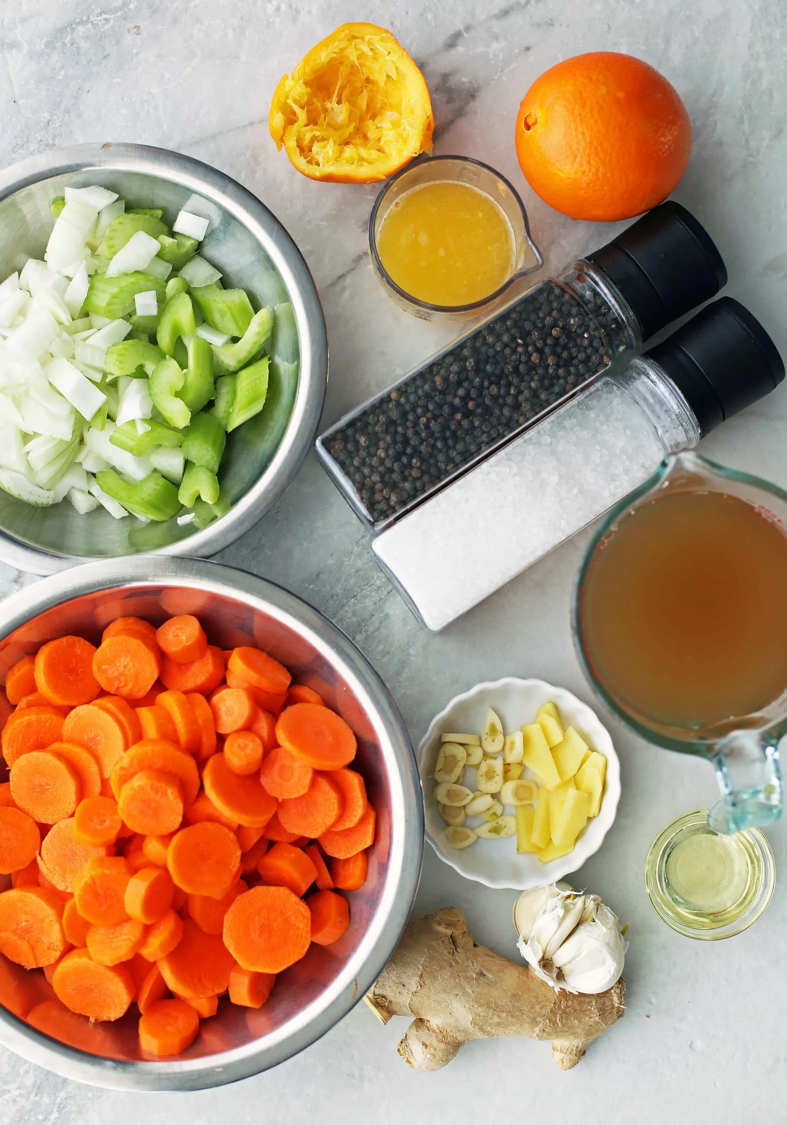 Bowls of carrots, onions, celery, ginger, garlic, vegetable broth, and orange juice along with salt and pepper shakers..