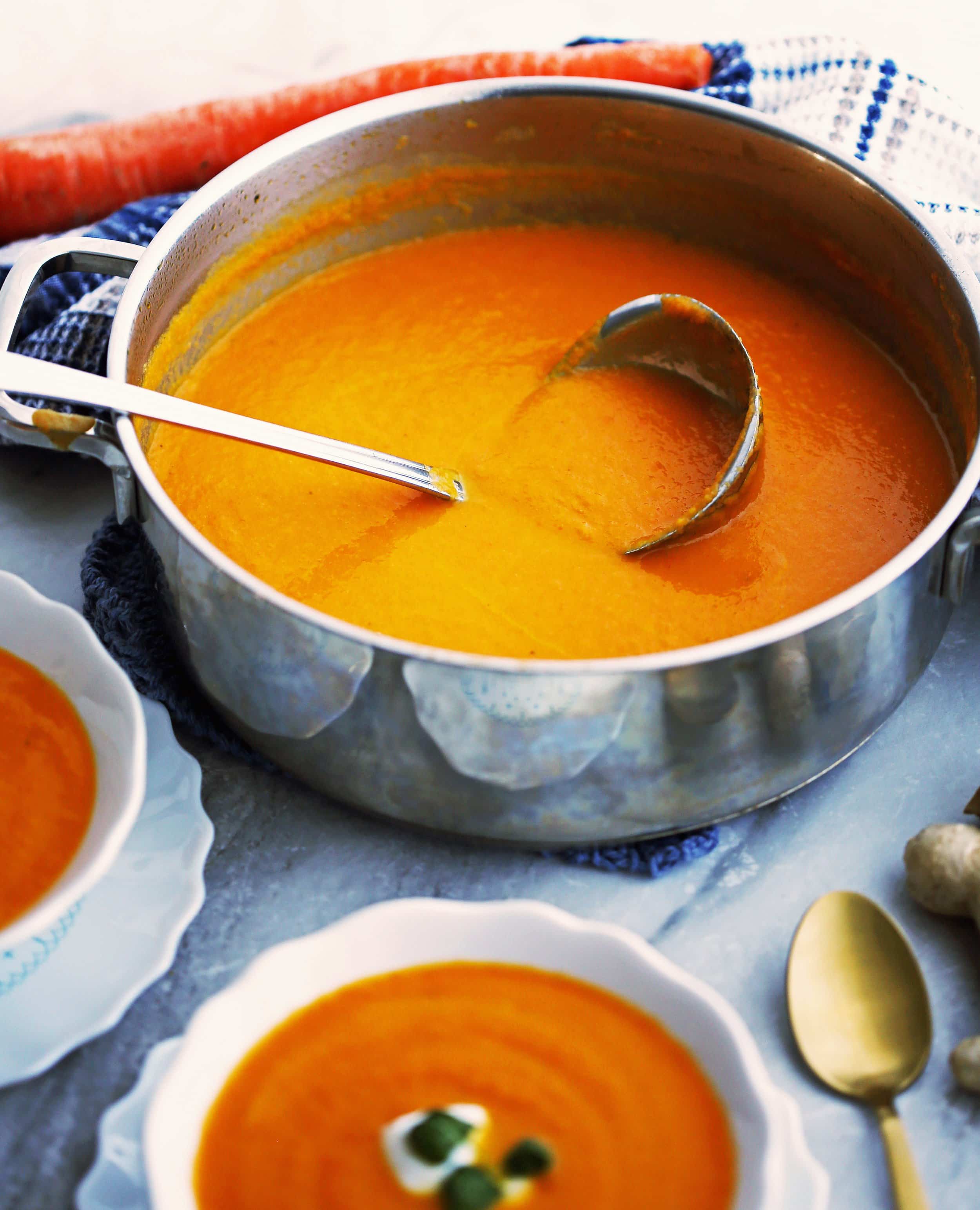 Healthy vegan carrot orange ginger soup in a large pot with ladle; bowls of soup around it.