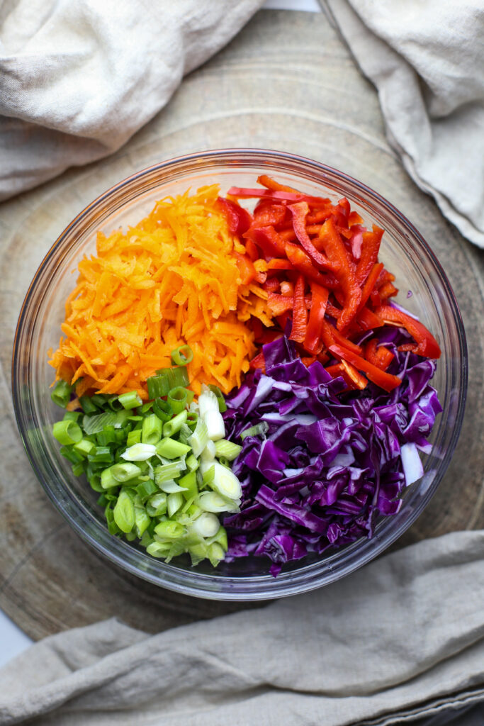 Overhead photo of sliced purple cabbage and red bell pepper, shredded carrots, and chopped green onions.