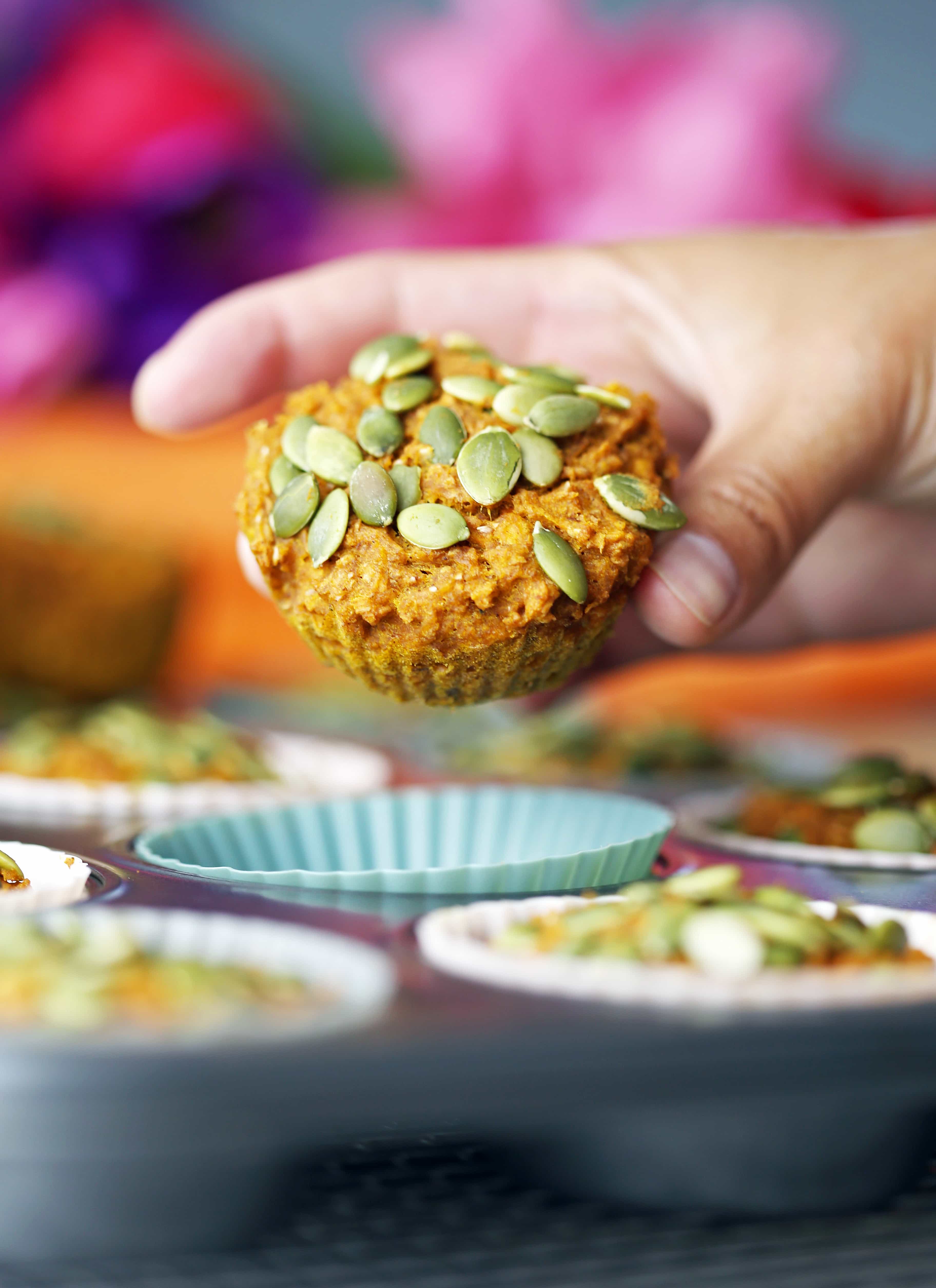 A hand holding up a carrot pumpkin muffin topped with pumpkin seeds.