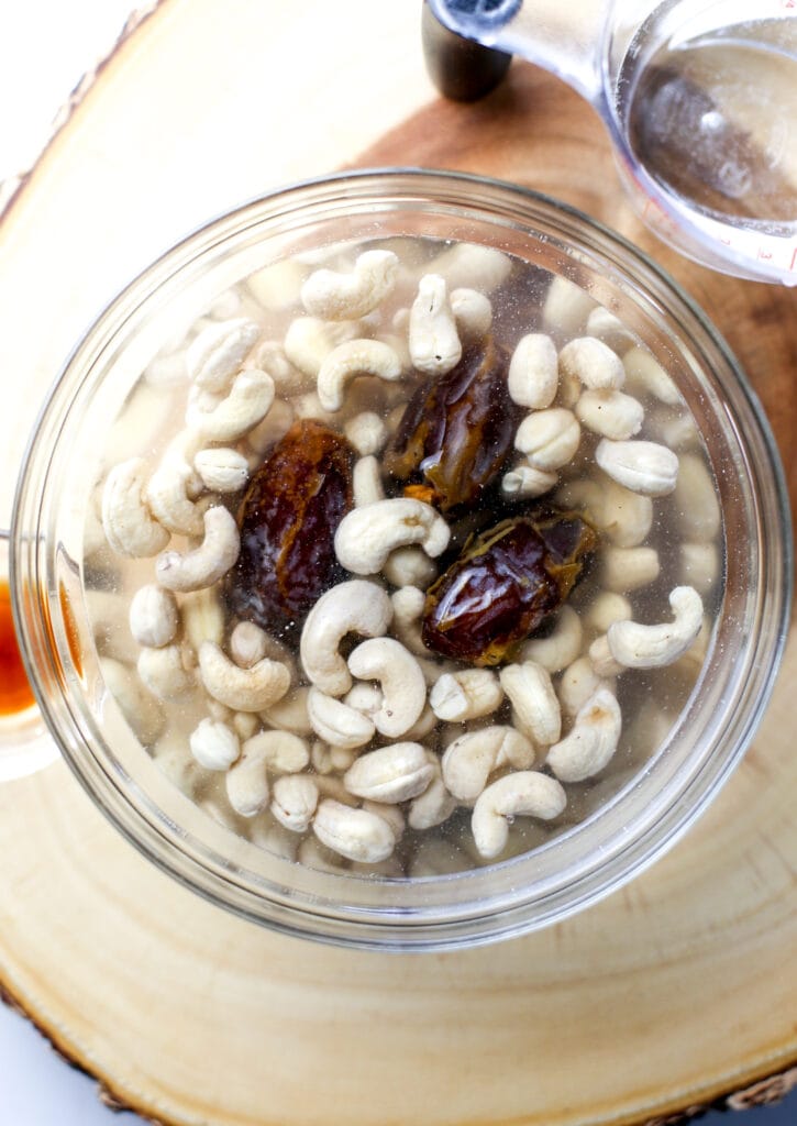 Overhead view of raw cashews and three Medjool dates covered in water in a glass bowl.