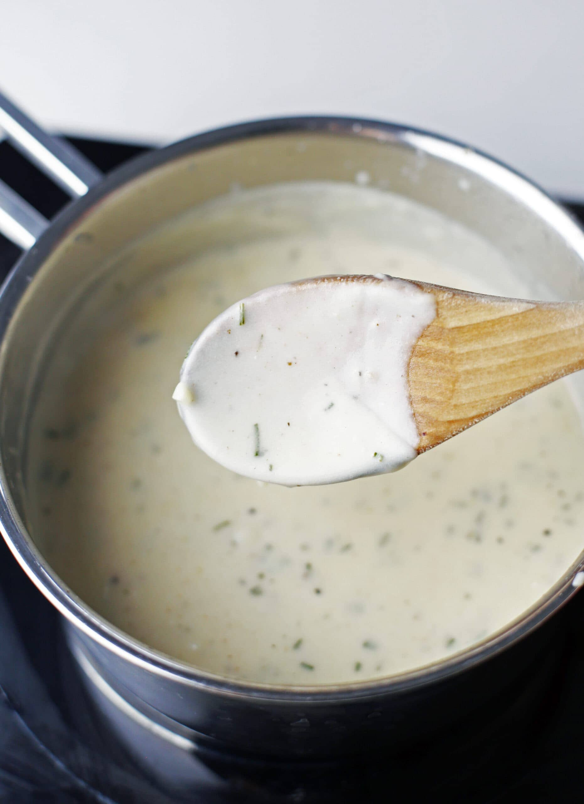 A wooden spoon with a coat of cheese sauce over saucepan of the same sauce.