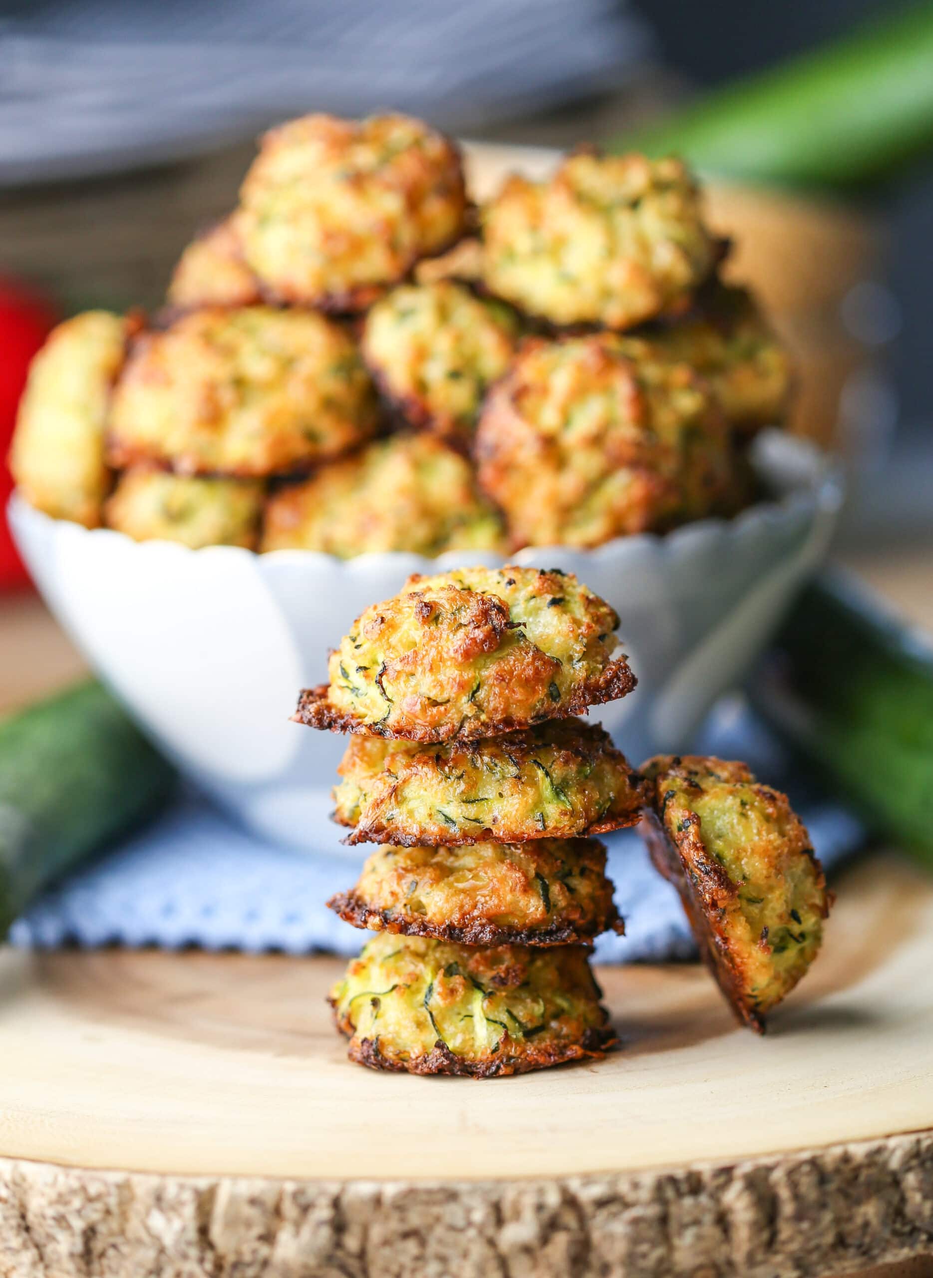 Four baked cheesy zucchini bites stacked on top of one another with more in a bowl behind them.
