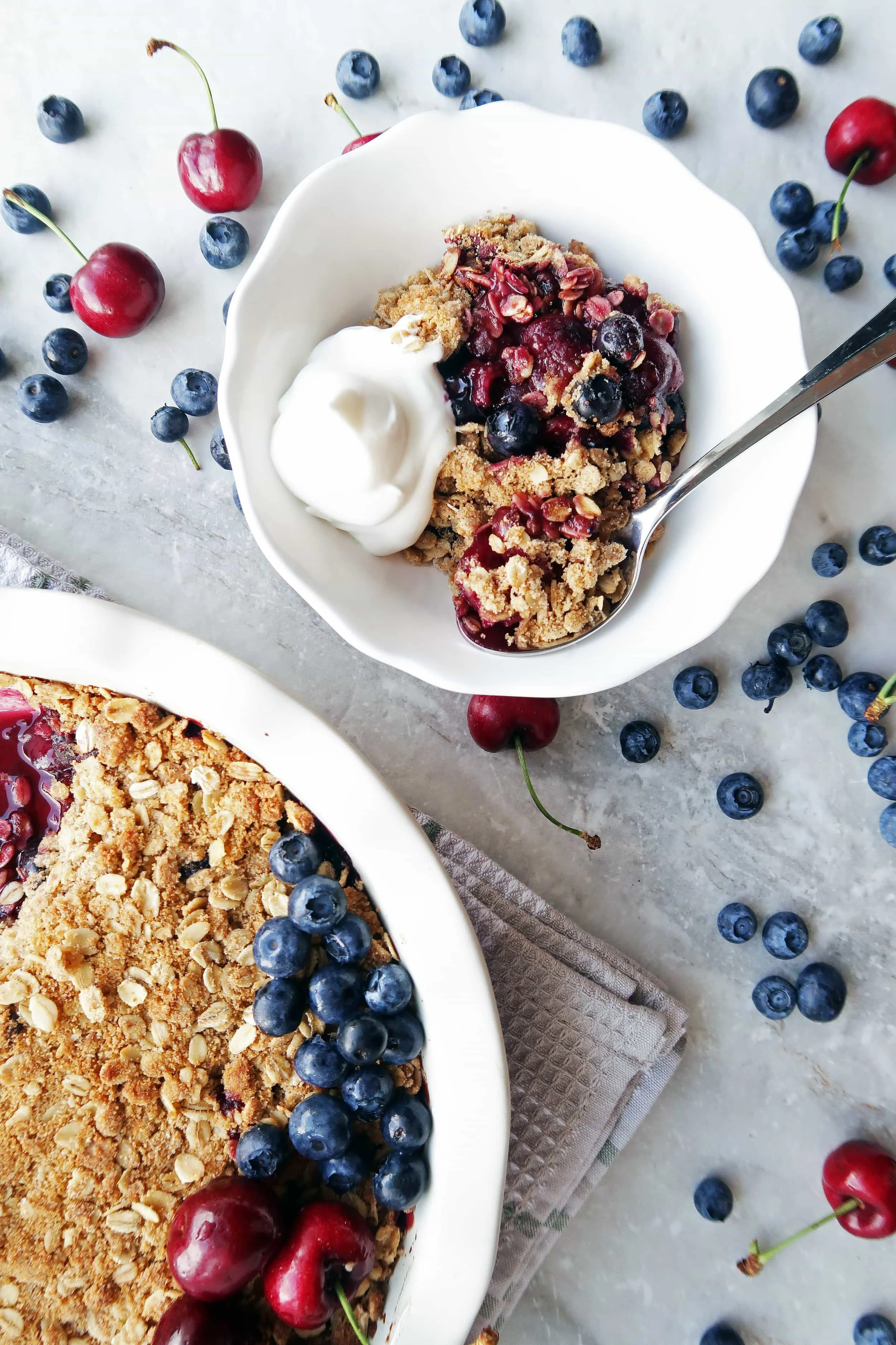 Cherry blueberry oat crisp with whipped cream in a white bowl with a spoon and a large pie dish of oat crisp.