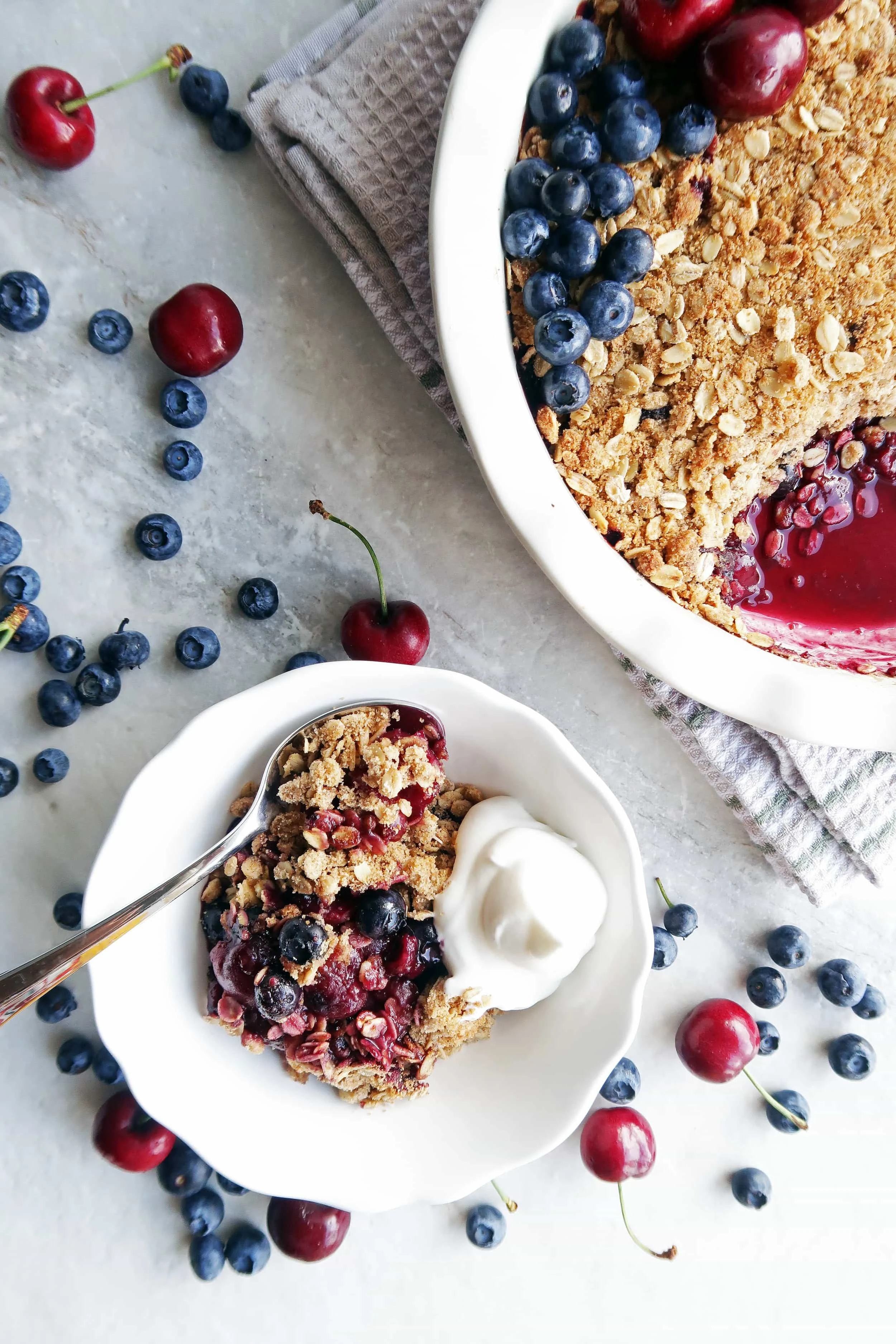 Cherry blueberry oat crisp with whipped cream in a white bowl with a spoon and oat crisp in a pie dish.