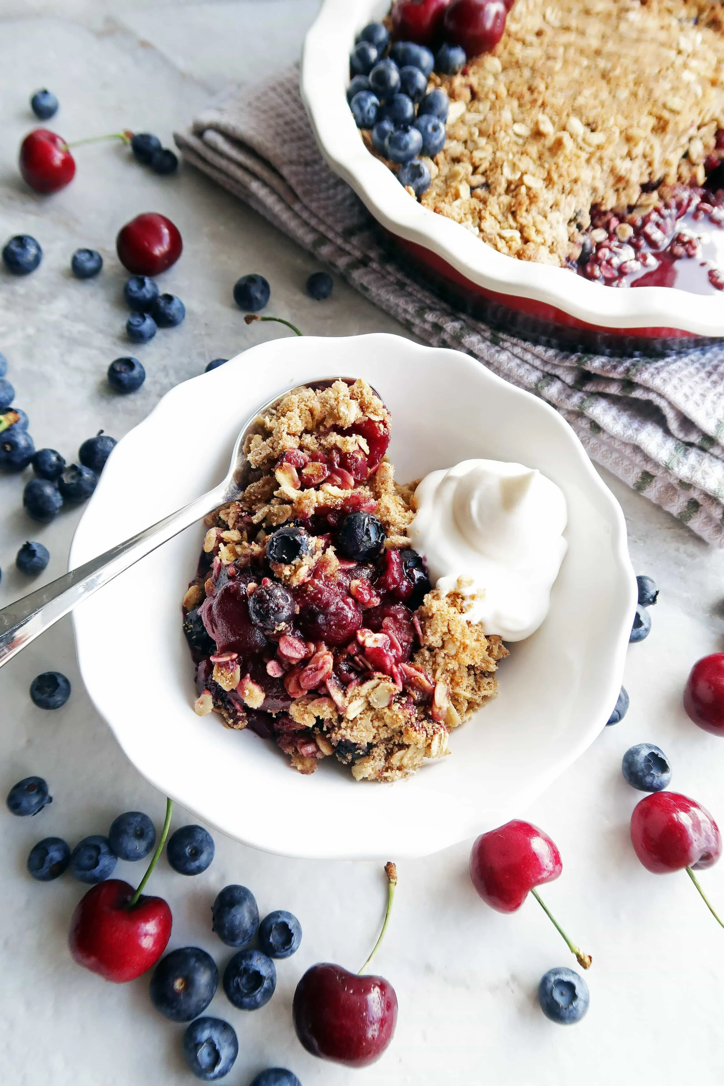 Cherry blueberry oat crisp with cream in a white bowl with a spoon and a pie dish of oat crisp in the background.