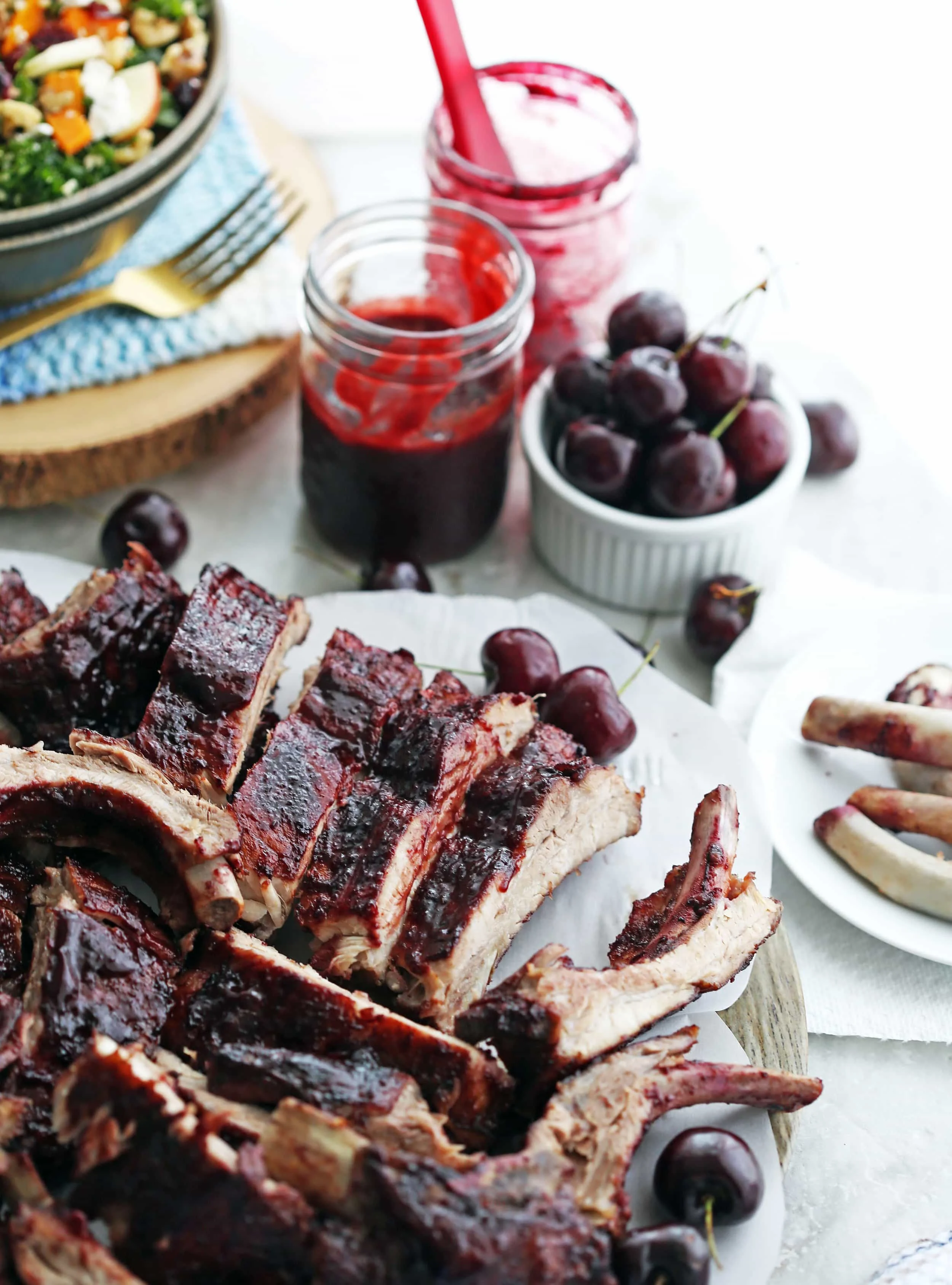 A platter of Baby Back Ribs covered with cherry chipotle sauce, a jar of sauce, and a bowl of fresh cherries on the side.