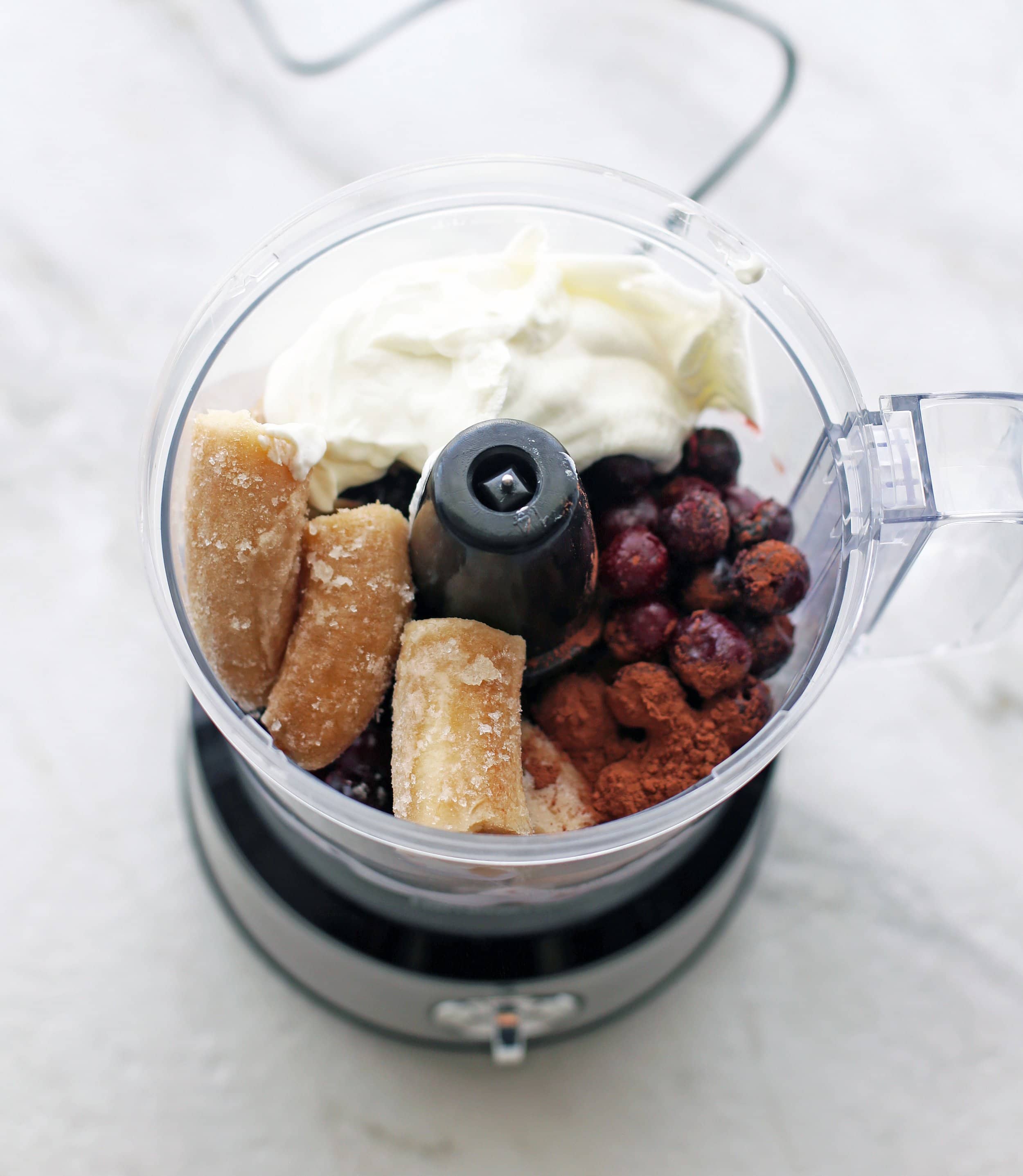 A food processor full of frozen cherries, frozen bananas, Greek yogurt, and cocoa poweder