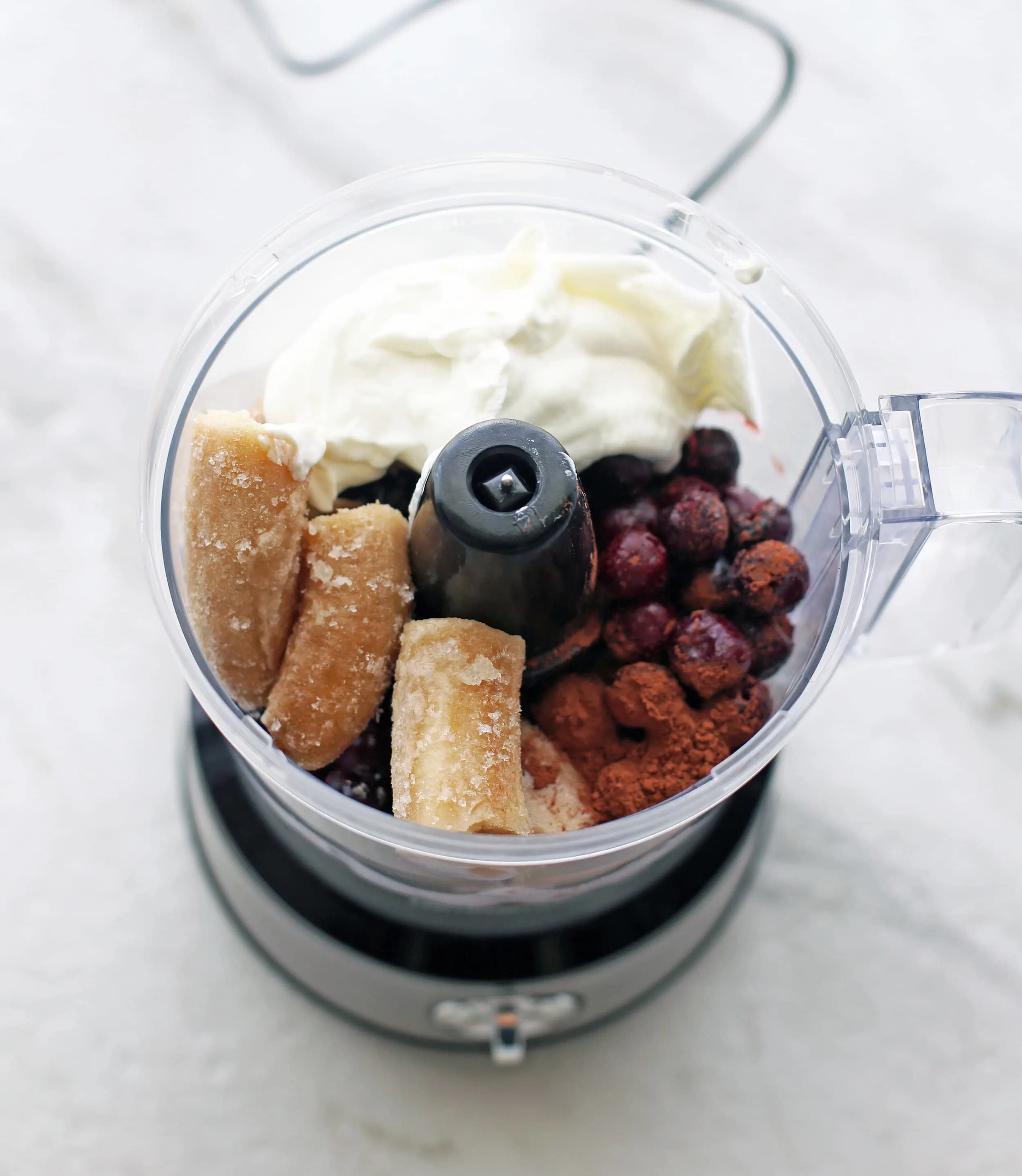 A food processor full of frozen cherries, frozen bananas, Greek yogurt, and cocoa poweder