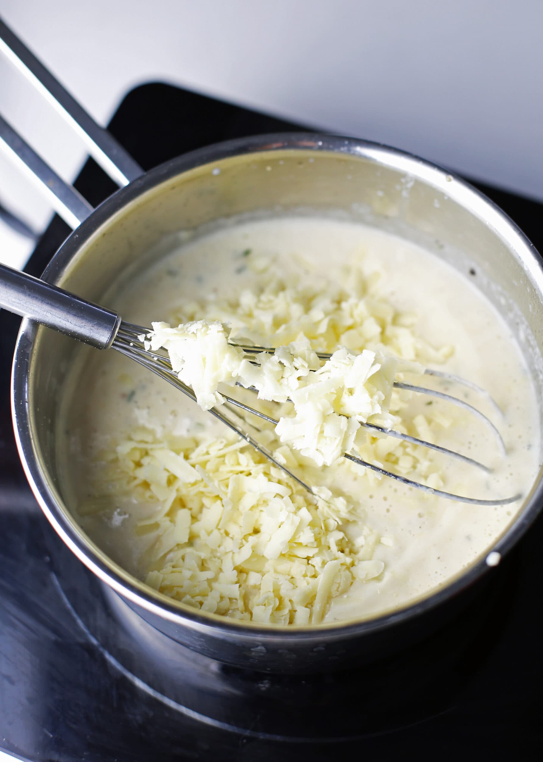 Grated cheddar cheese just added to white sauce in a saucepan with a whisk.