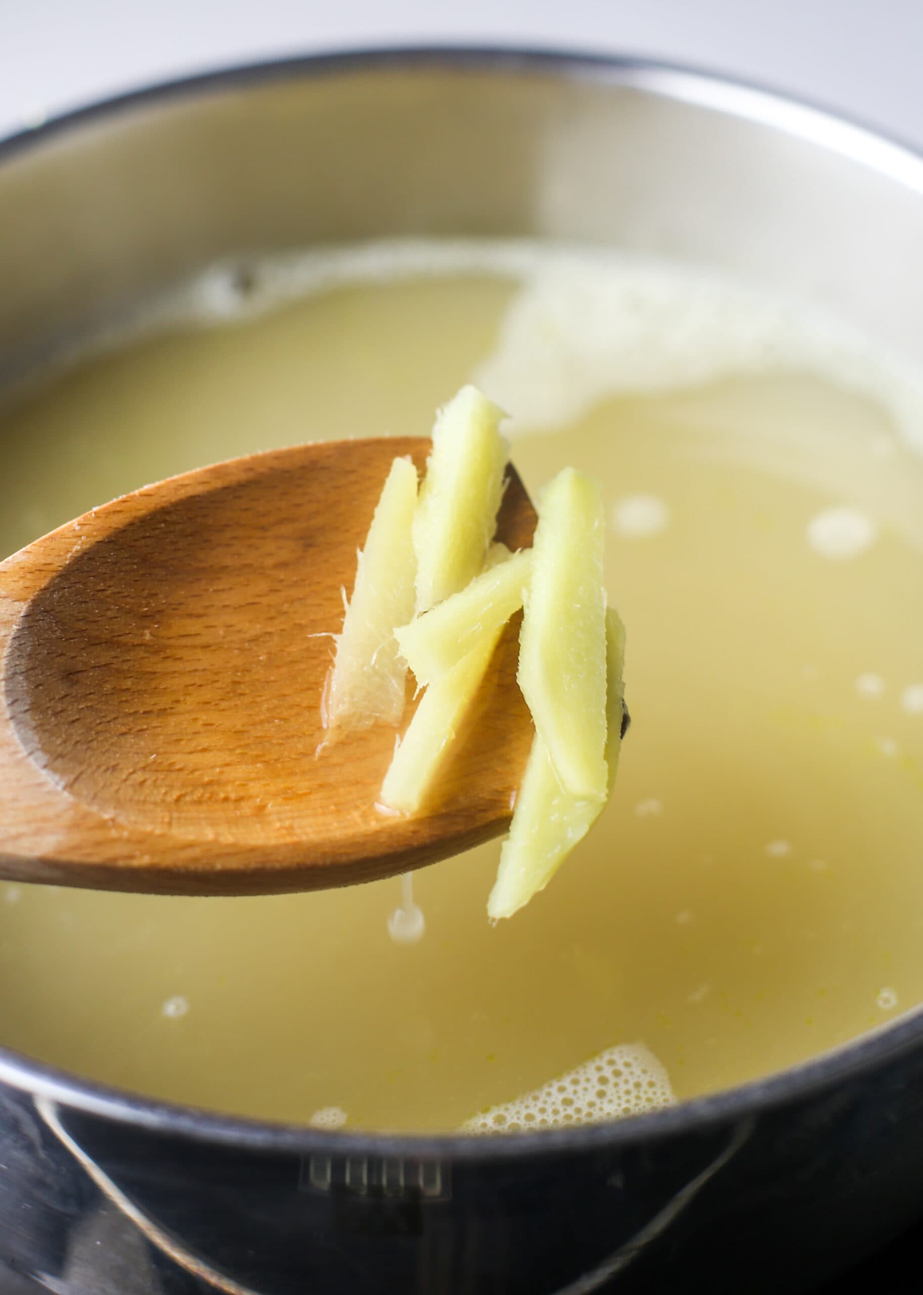 A wooden spoon holding ginger slices over a pot of ginger-flavoured chicken broth.