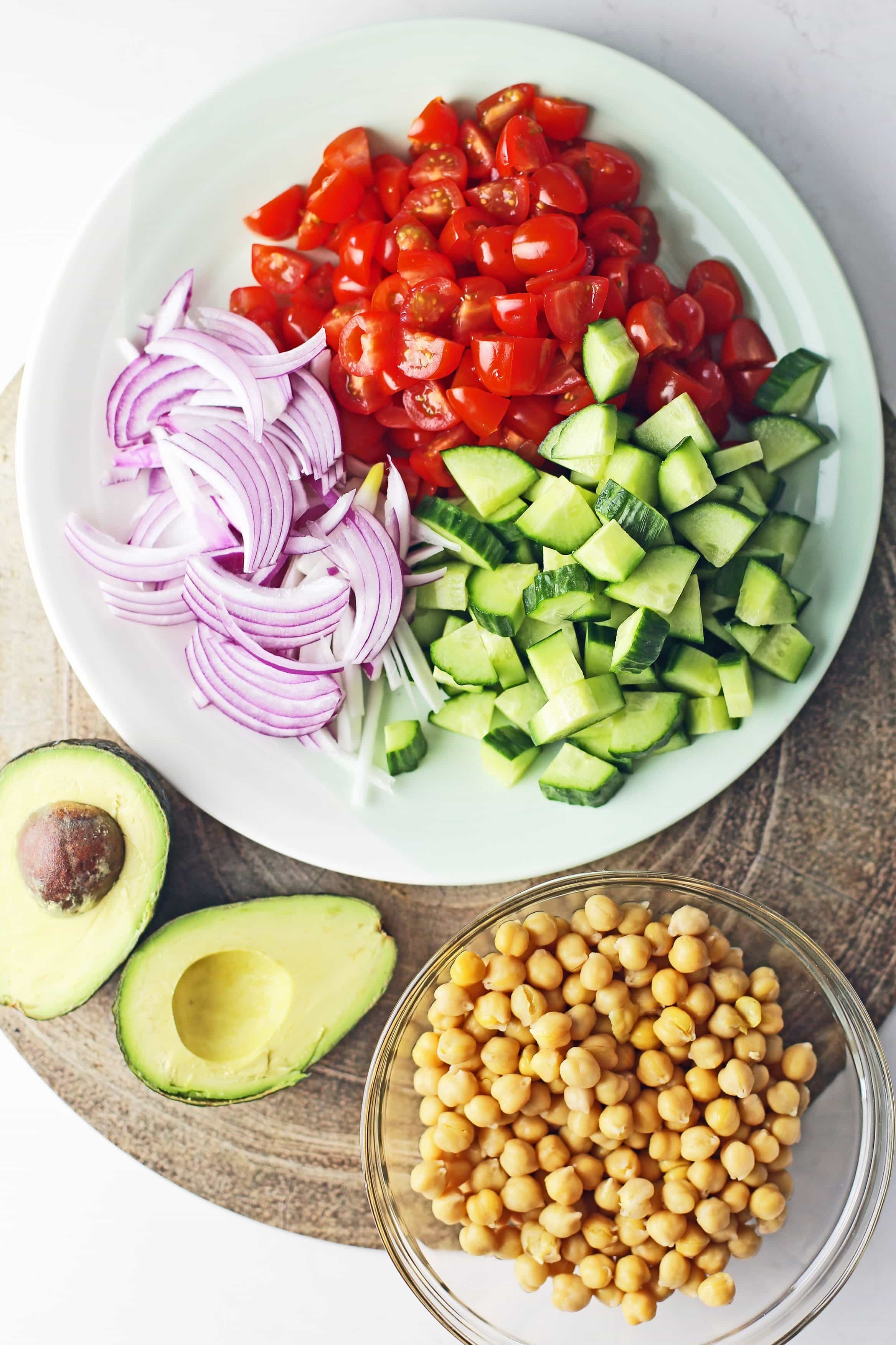 A large plate containing chopped and sliced English cucumber, grape tomatoes, and red onions, a bowl of cooked chickpeas, and an avocado cut in half.