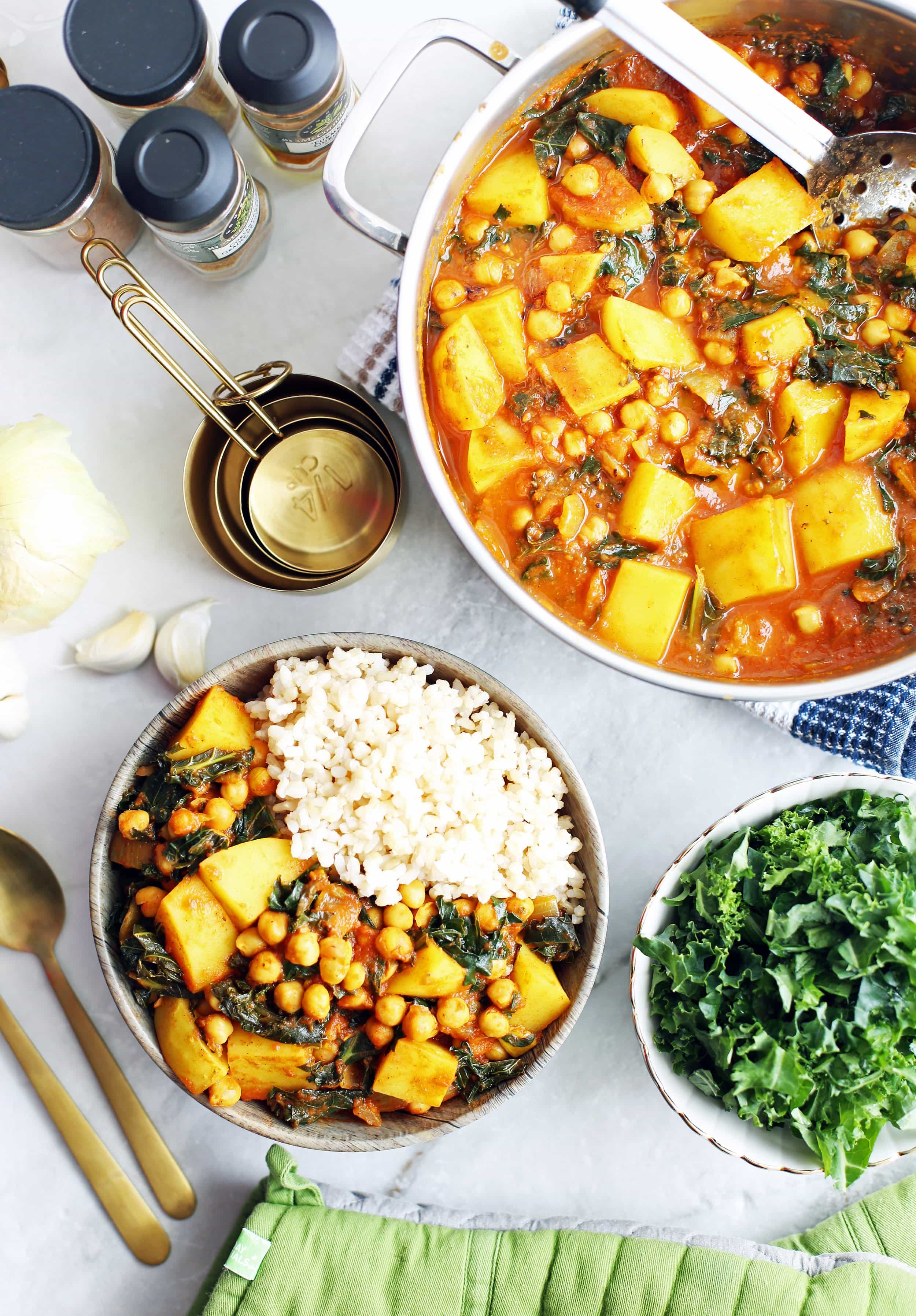 An overhead view of a bowl of chickpea kale and potato curry, a pot of curry, and a bowl of raw kale.