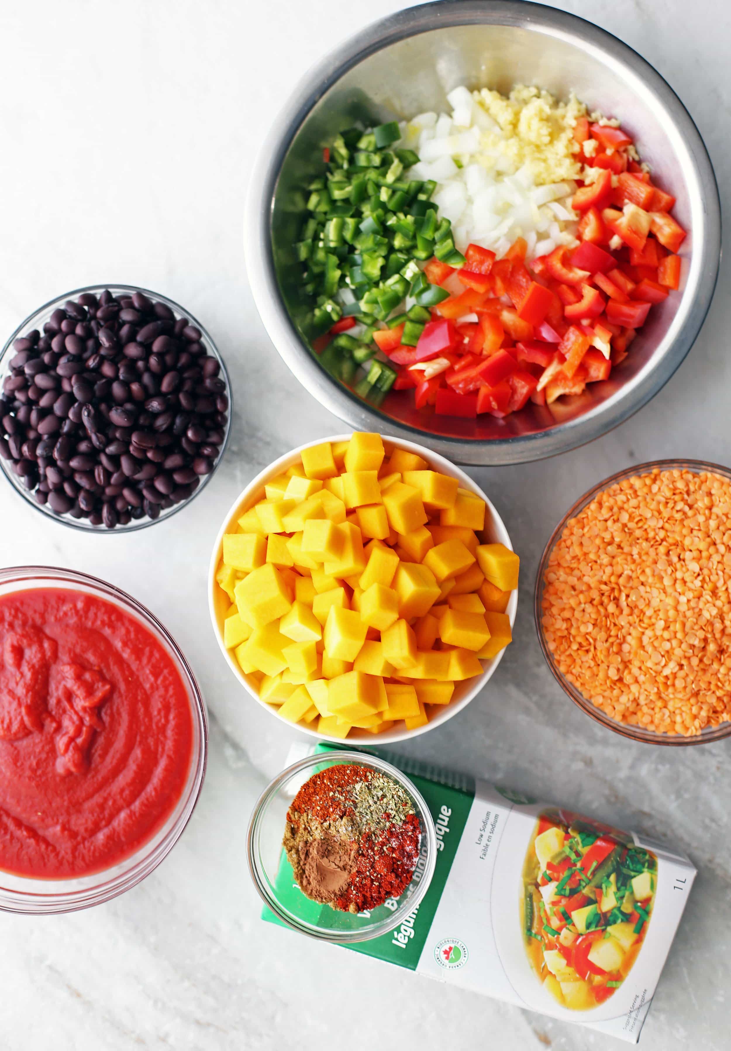 Chopped vegetables, vegetable broth,with bowls of black bean, lentils, crushed tomatoes, and spices.