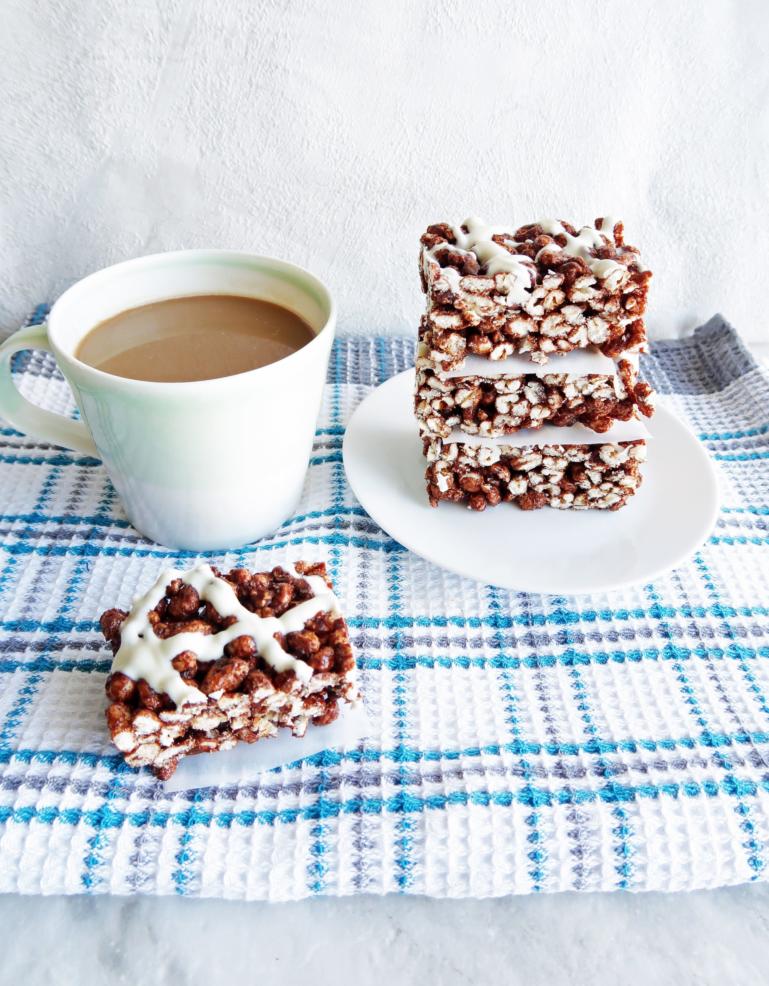 Three Chocolate Marshmallow Puffed Wheat Squares stacked on a white plate along with another puffed wheat square and cup of coffee beside the plate.
