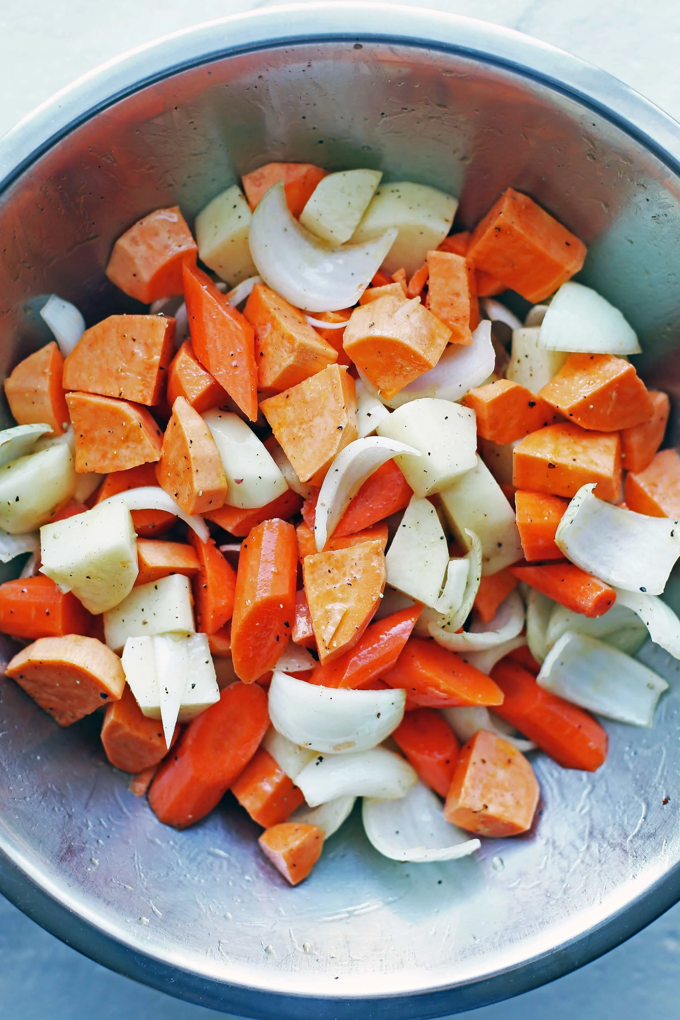 Chopped carrots, potatoes, sweet potatoes, and onions in a large stainless steel bowl.