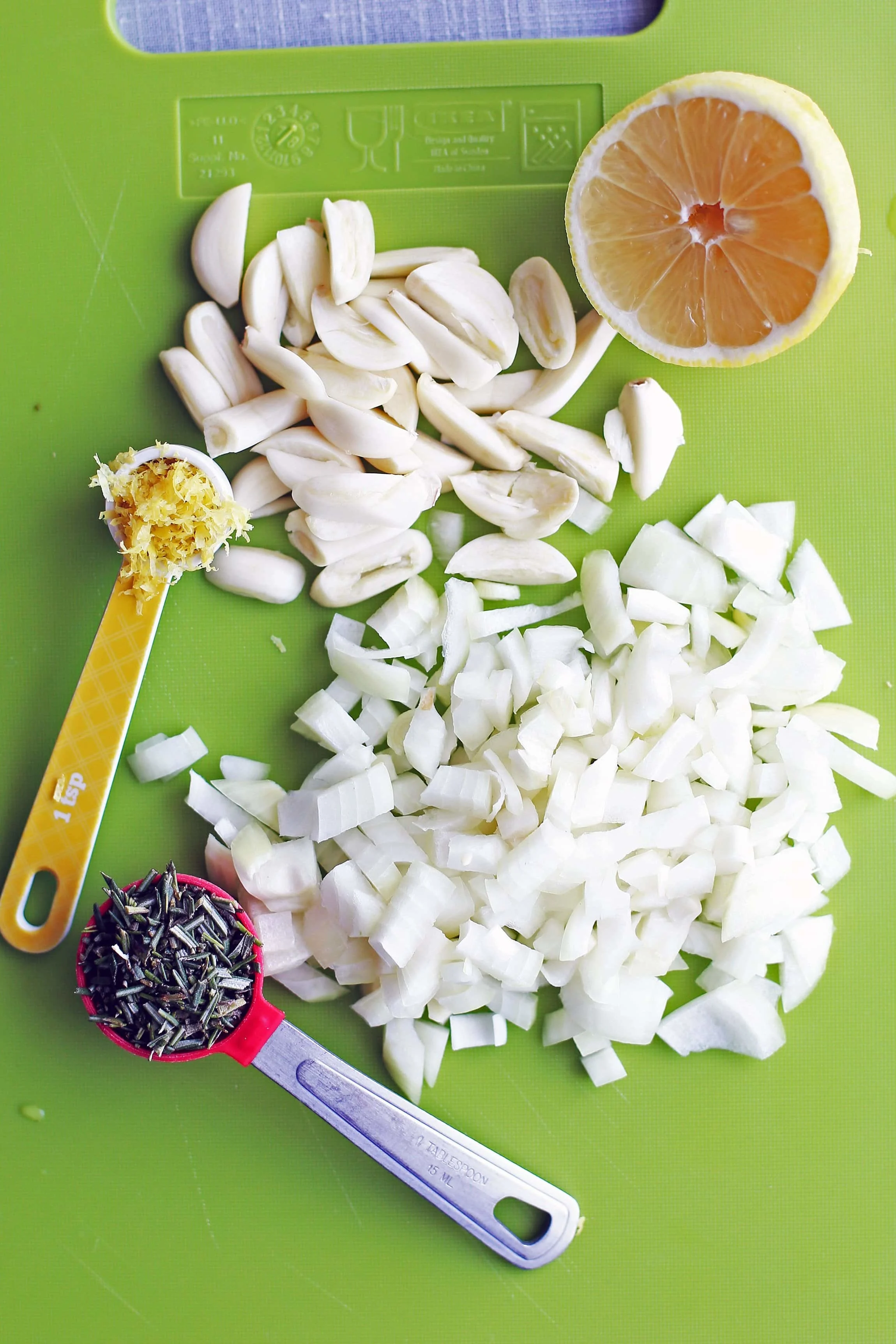 Diced onions, quartered garlic cloves, lemon zest, half lemon, and minced fresh rosemary on a green cutting board.