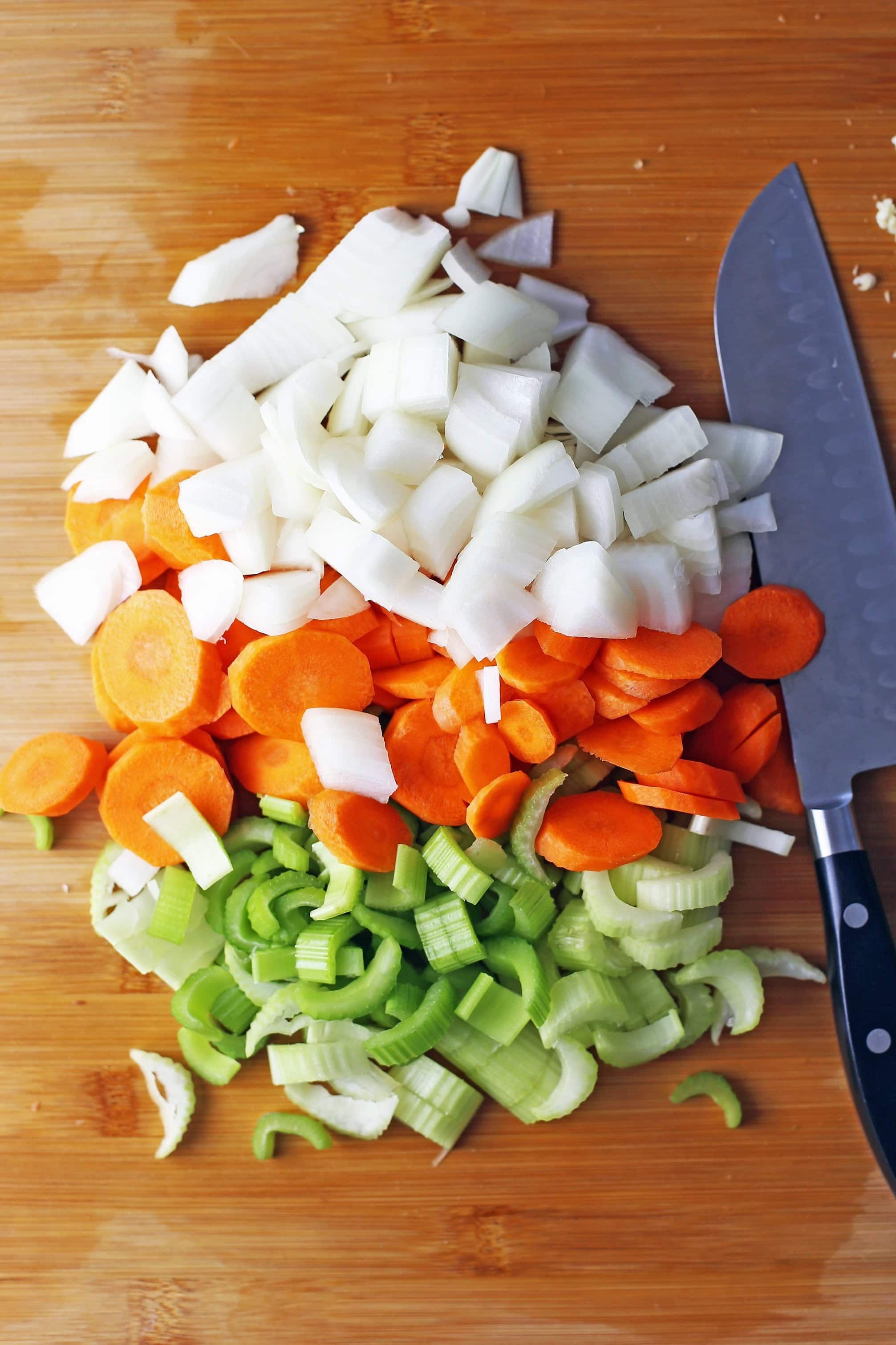 Chopped white onion, carrots, and celery on wooden cutting board with a knife on the the side of the vegetables.