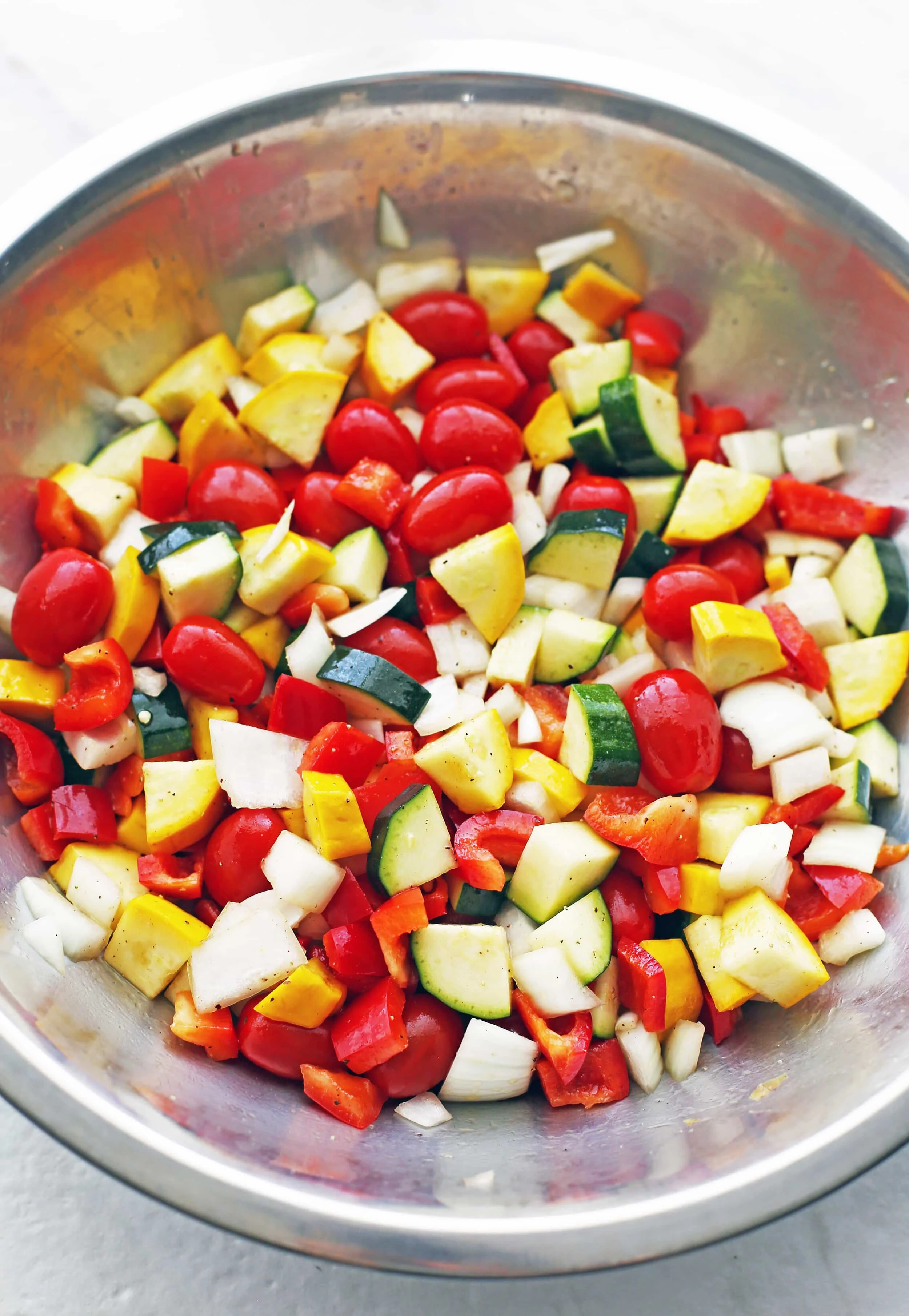 A large metal bowl containing grape tomatoes and chopped zucchini, yellow squash, bell peppers, and onions.