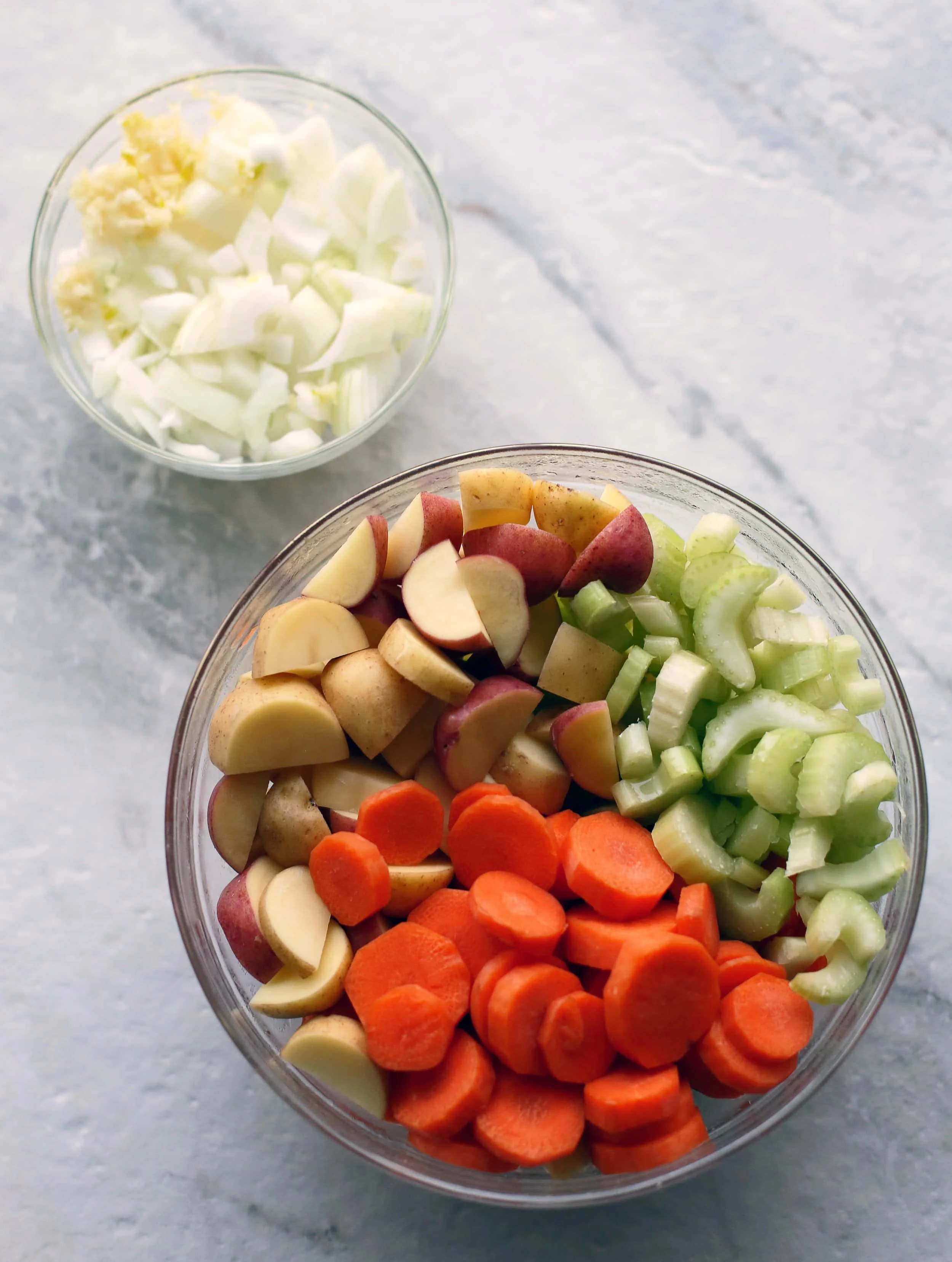 Two glass bowls filled with garlic, onions, carrots, potatoes, and celery.