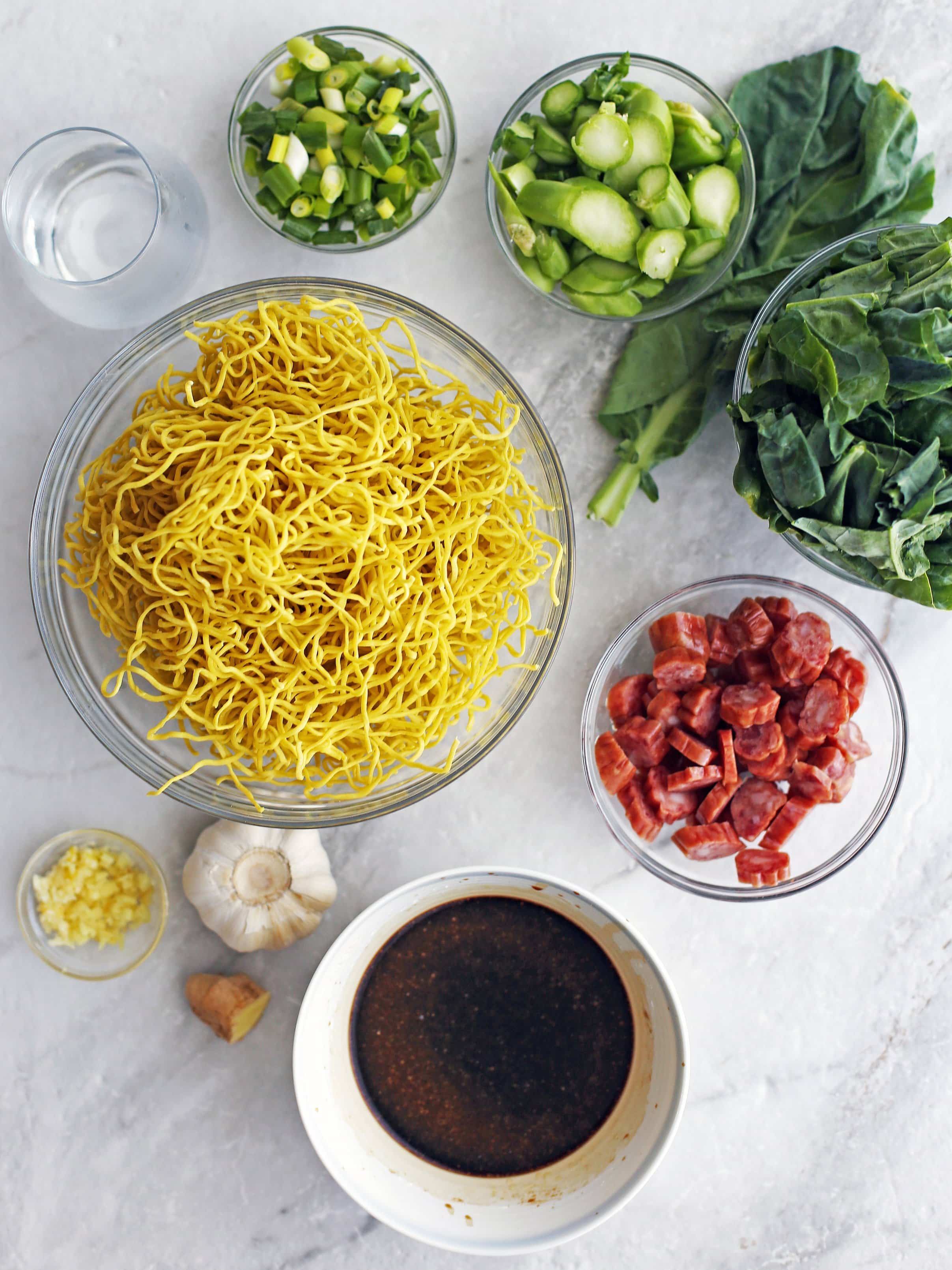 Overhead view of stir-fry sauce, chow mein noodles, Chinese sausage, ginger, garlic, gai lan, green onions, and water.