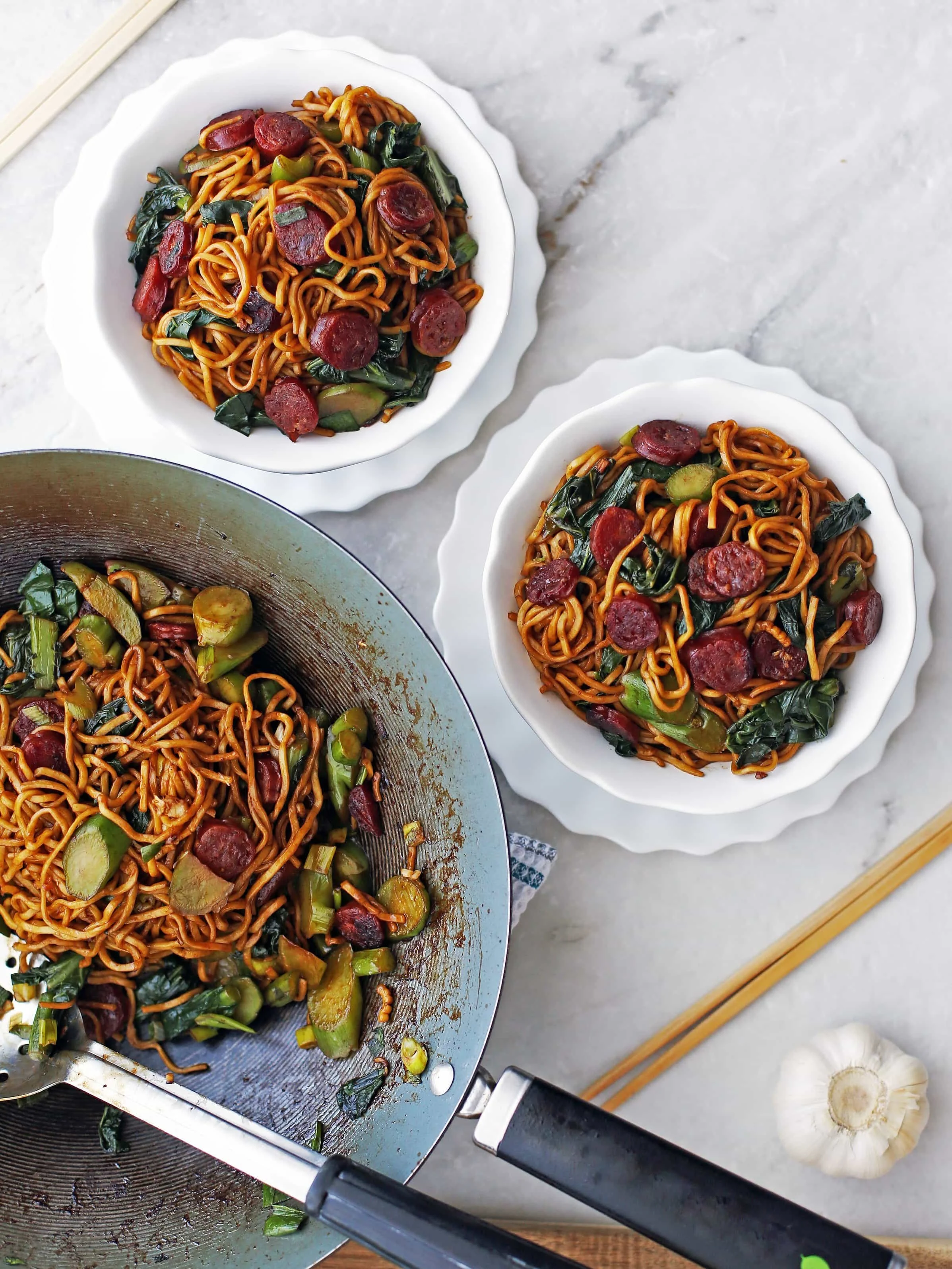 Two bowls of Lo mein noodles with Chinese sausage and gai lan and more in a large wok.