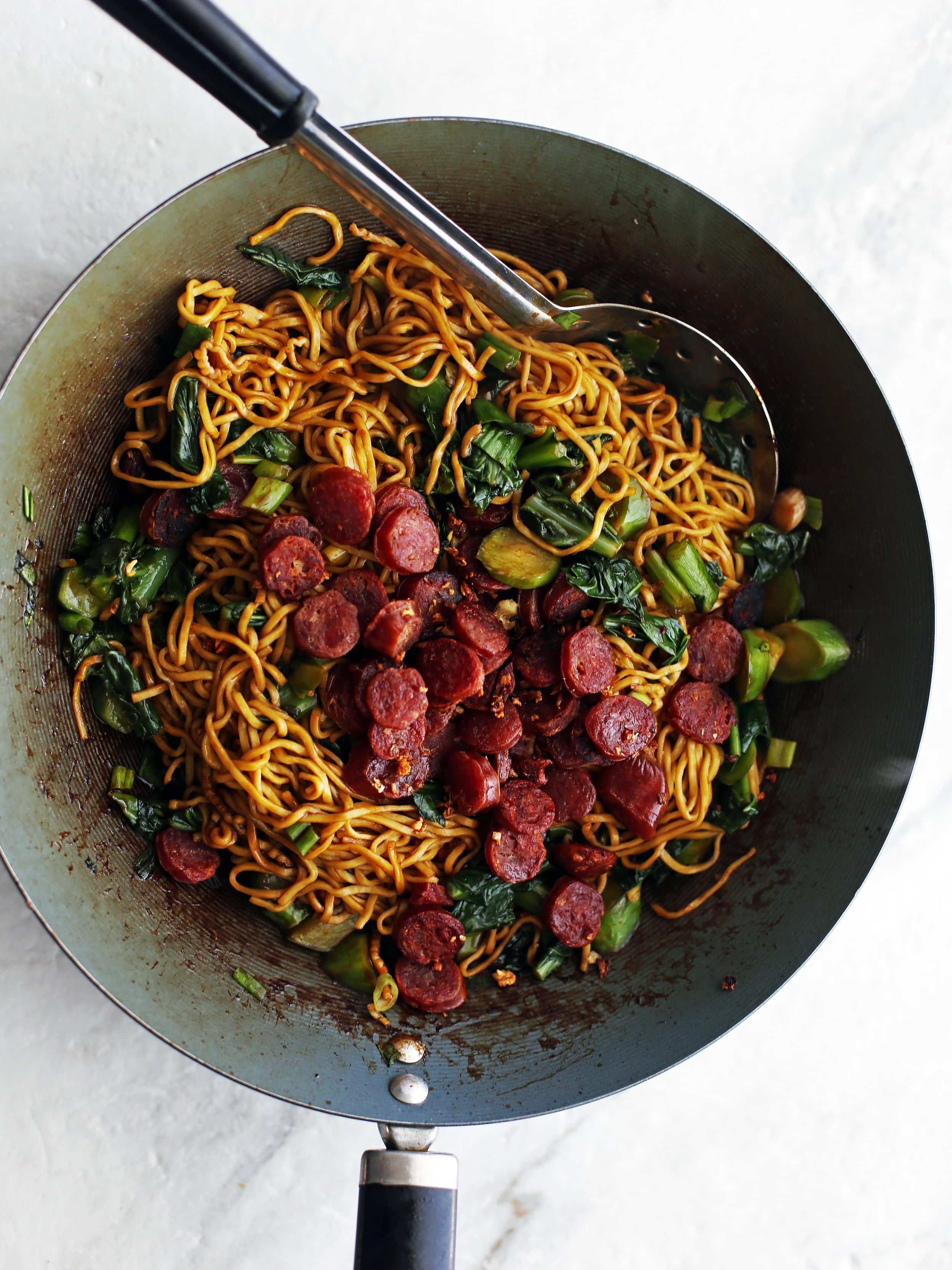 Lo mein noodles with Chinese sausage (lap cheong) and gai lan (Chinese broccoli) in a large wok.