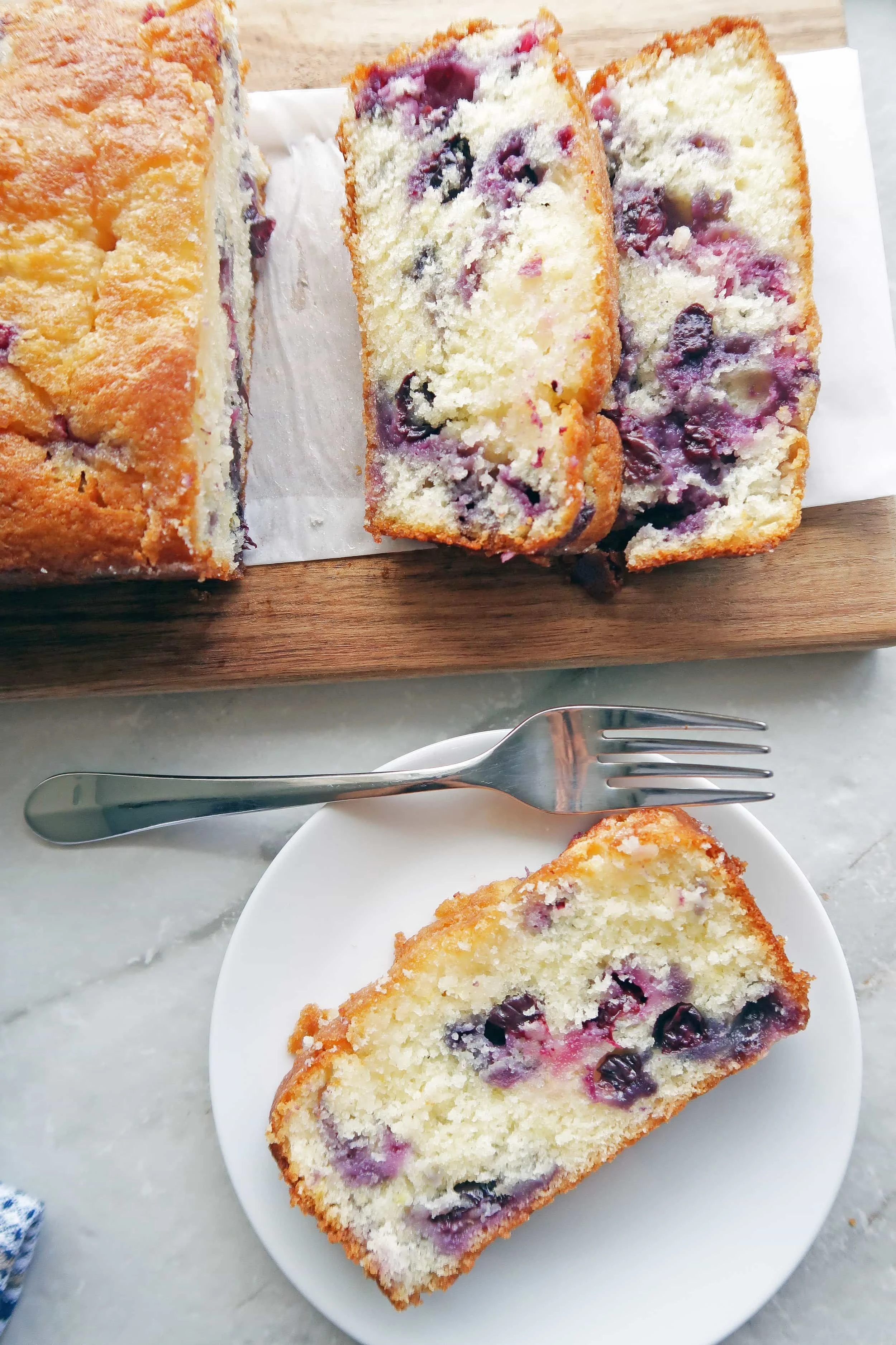 A slice of fluffy Classic Lemon Blueberry Loaf Cake on a white plate; the remaining sliced loaf cake on a long wooden board beside it.