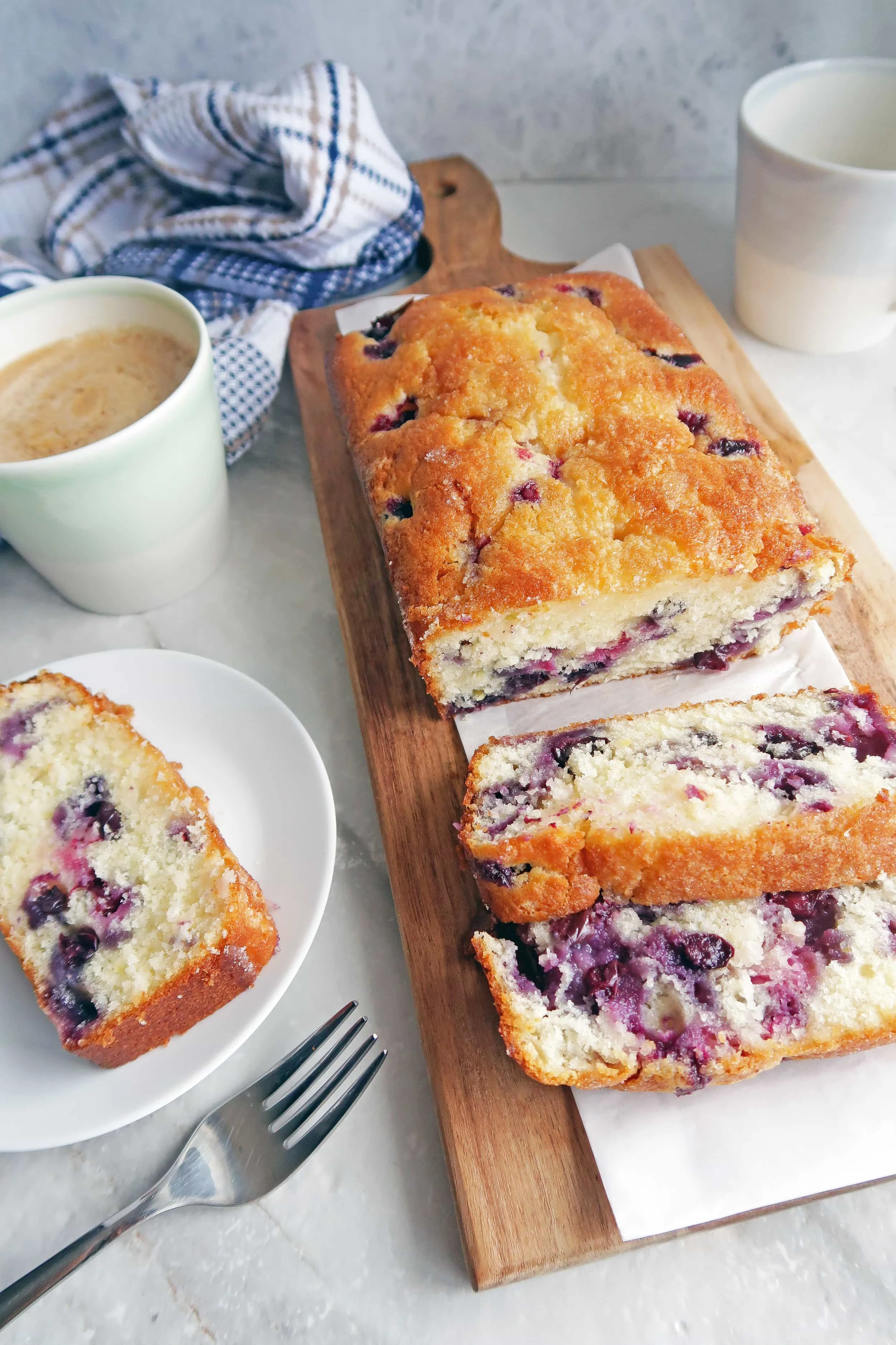 A slice of fluffy Classic Lemon Blueberry Loaf Cake on a white plate; the remaining loaf cake on a long wooden board.