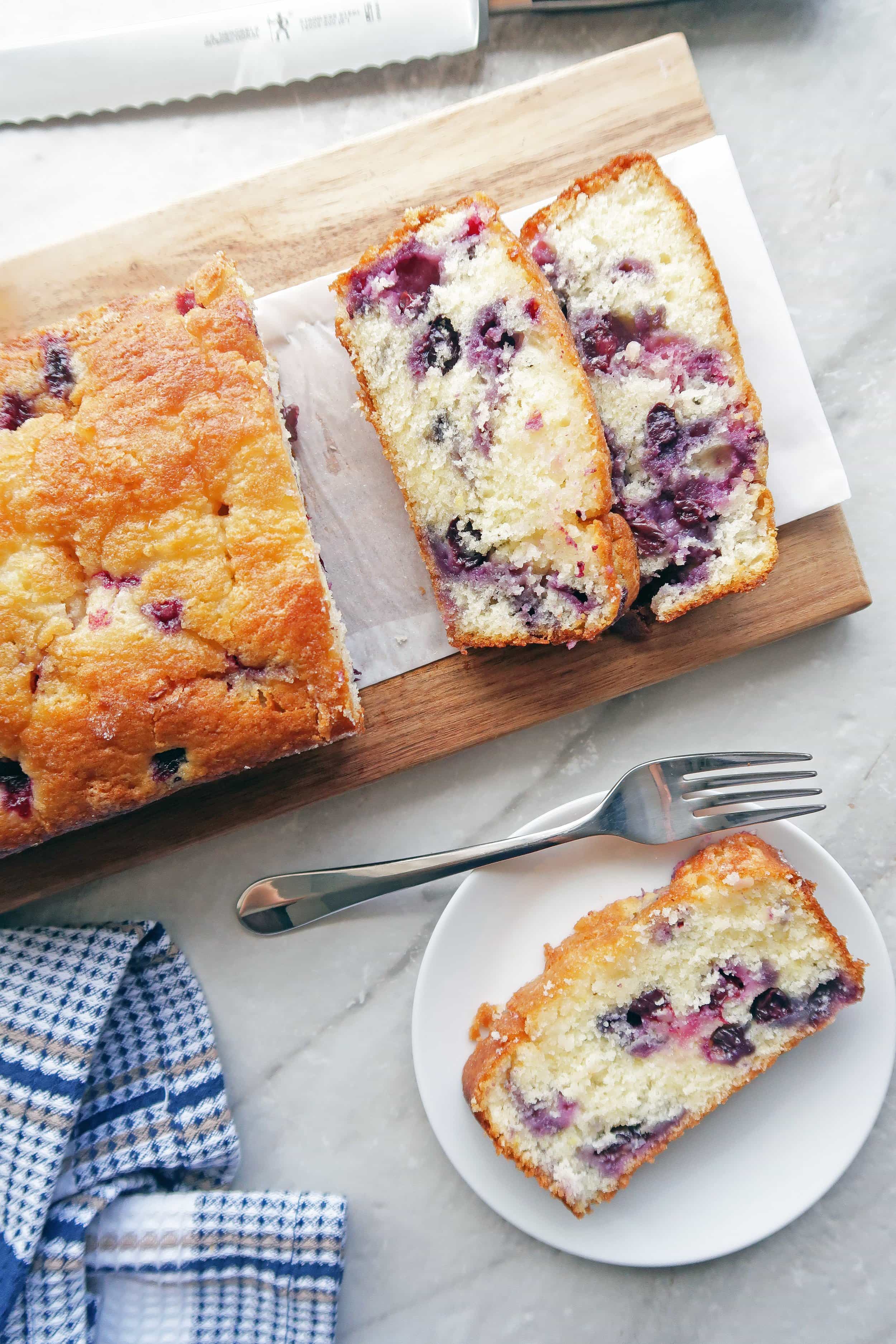 Piece of Classic Lemon Blueberry Loaf Cake on a white plate with fork; the remaining loaf cake on a long wooden board.