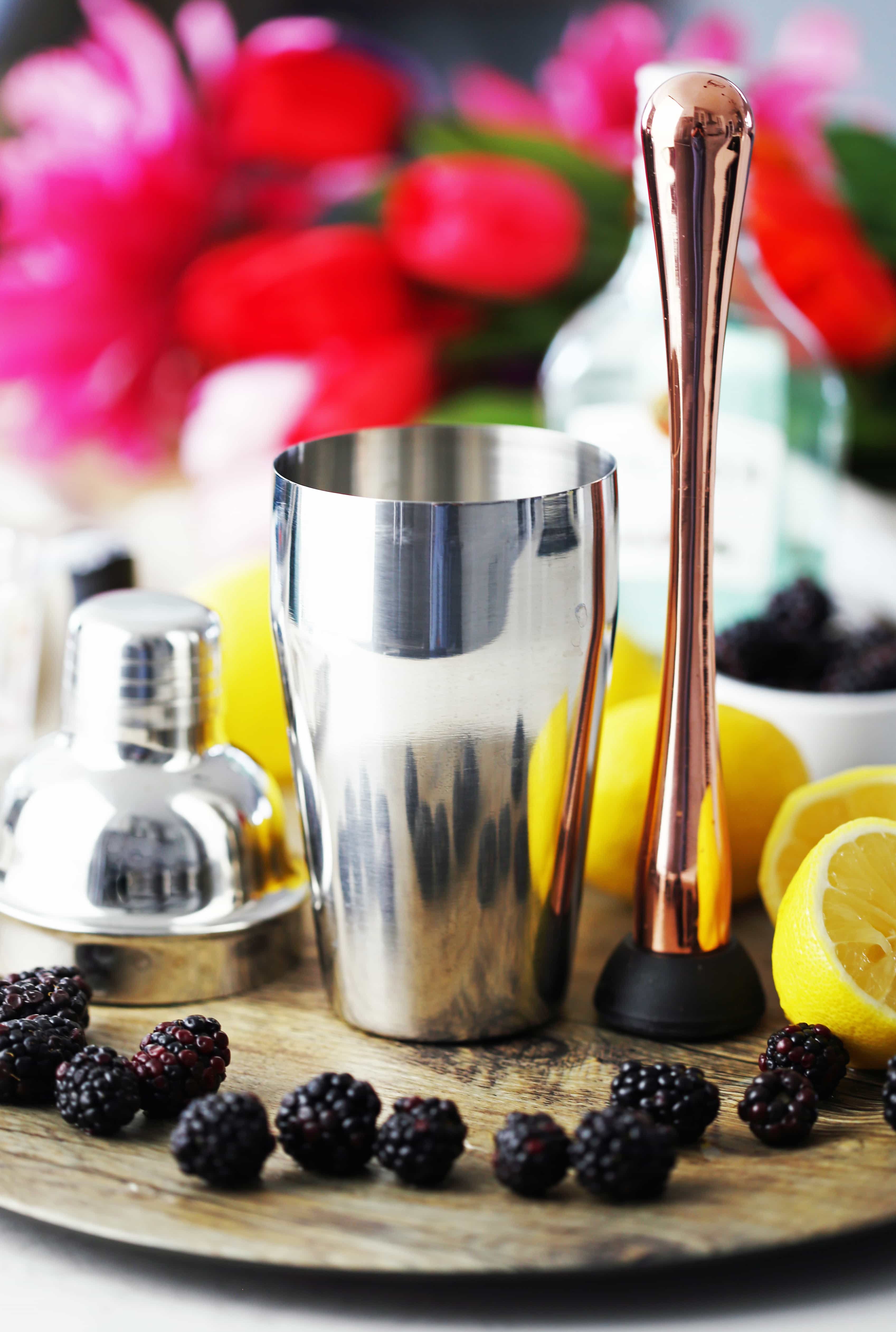 A stainless steel cocktail shaker and rose gold muddler on a wooden platter with blackberries and lemons