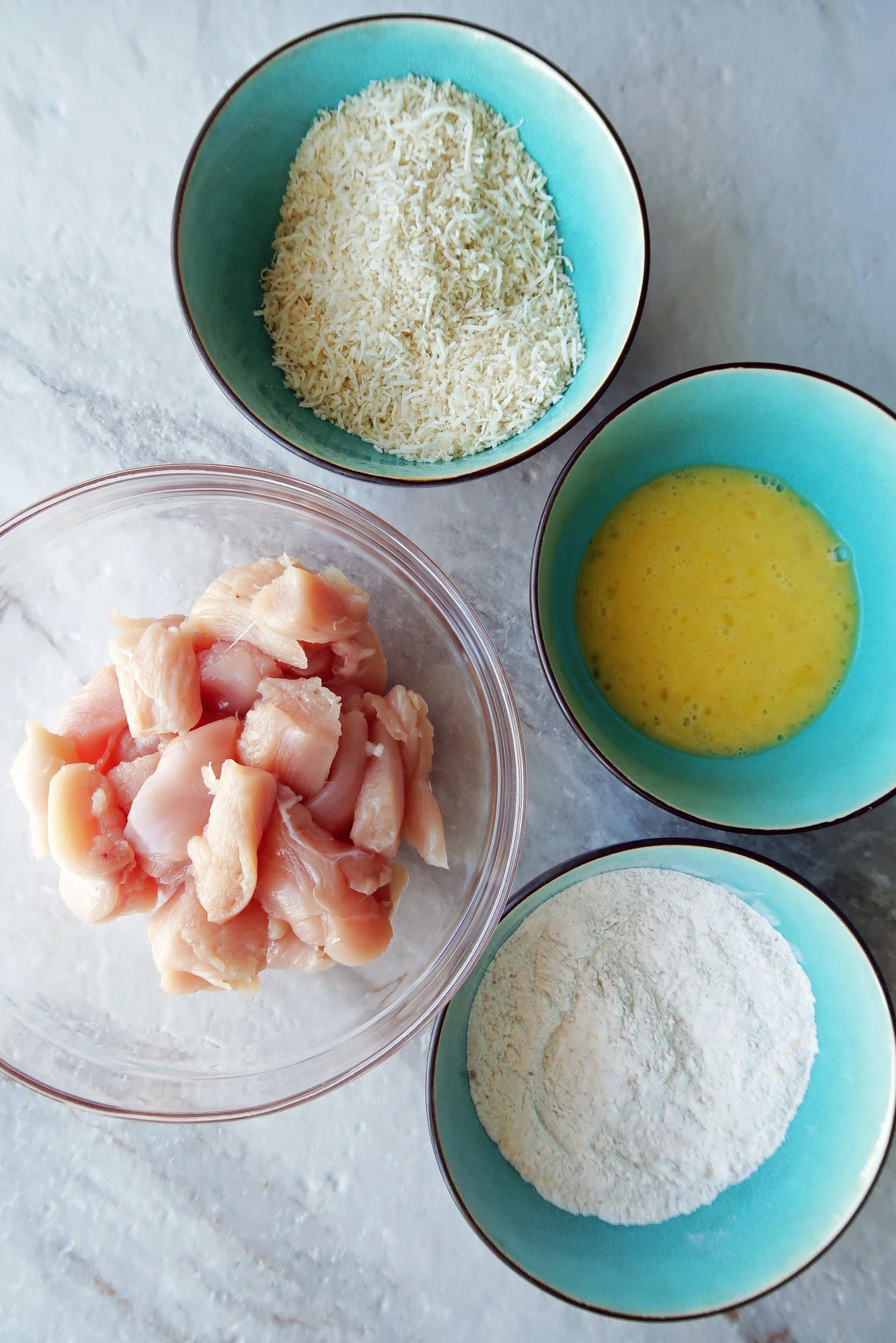 Four bowls for dredging: flour with spices, eggs, shredded coconut with Panko breadcrumbs, and raw chicken pieces.